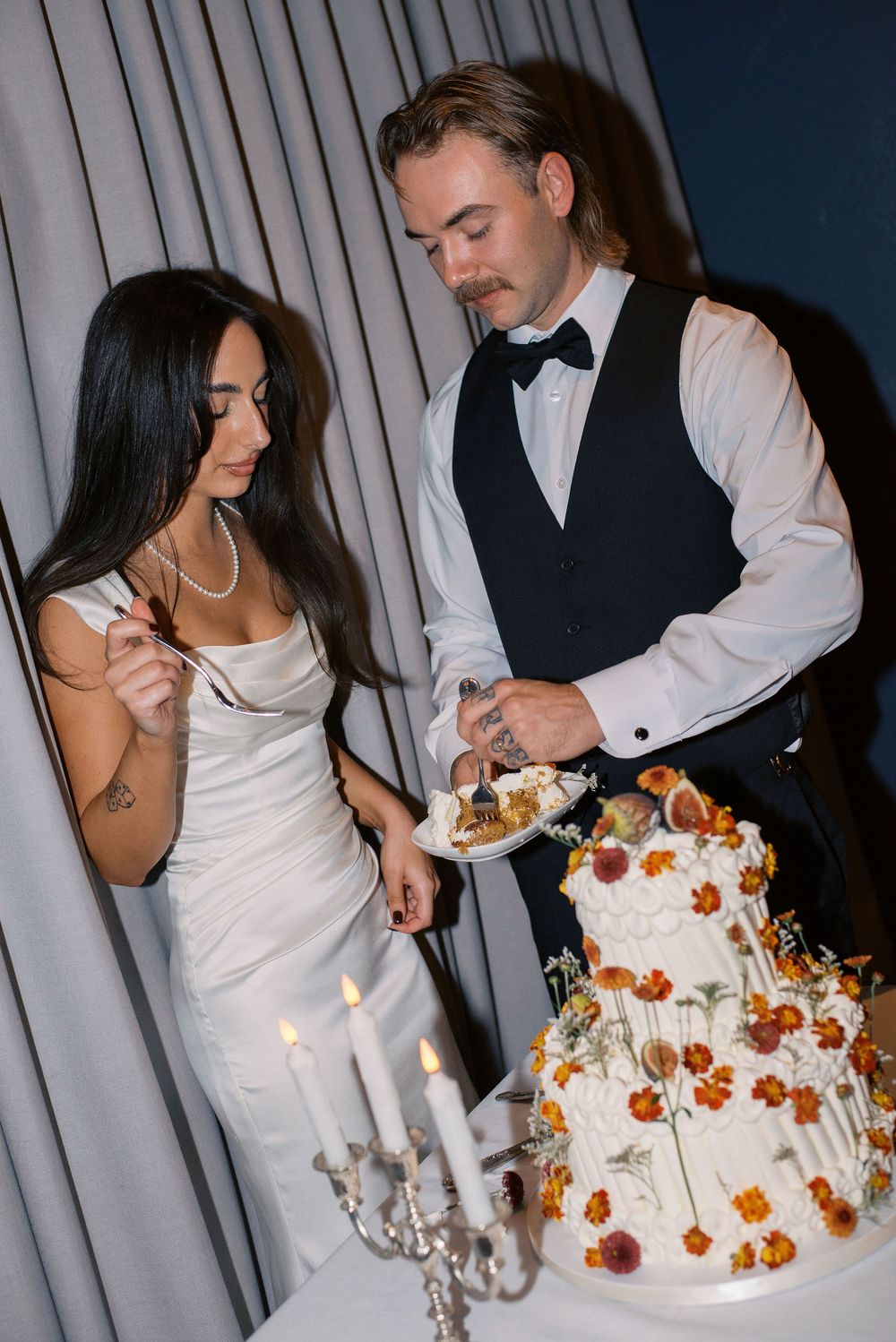 A wedding couple cuts into their elegant white tiered cake decorated with orange flowers and lit candles.