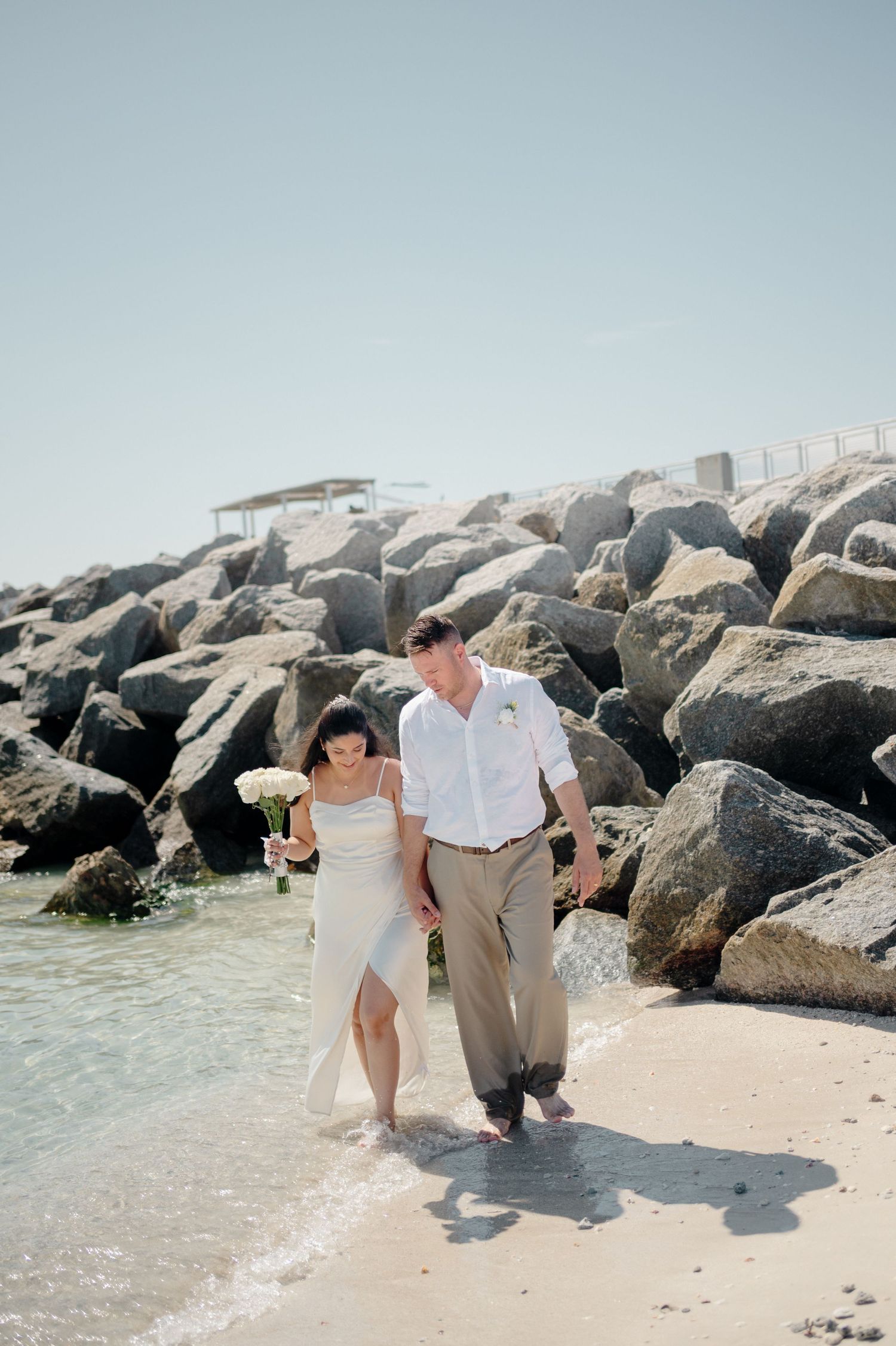 Romantic couple sitting together on rocky beach jetty by the ocean in casual beach wedding attire.