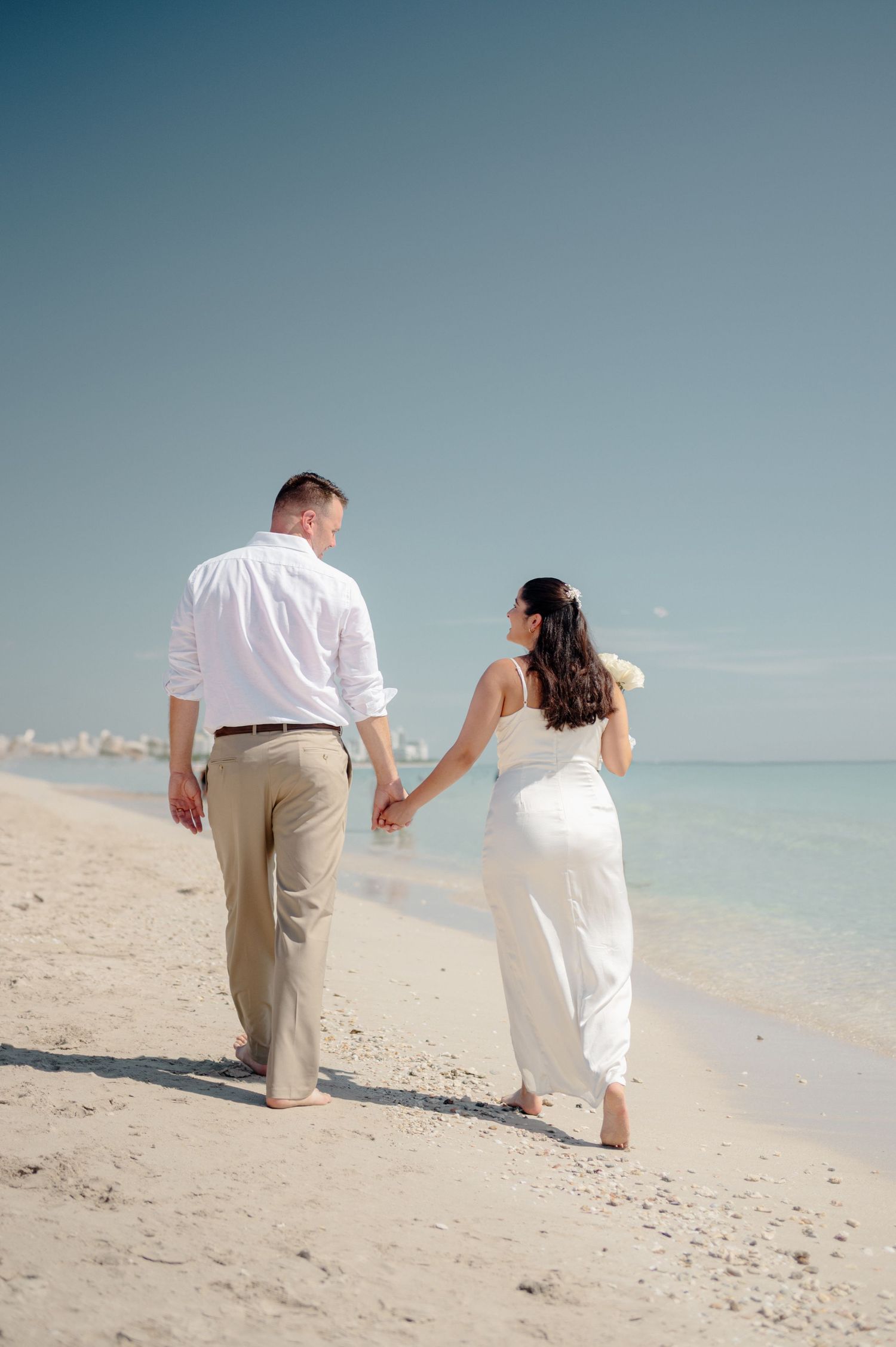 A couple in white clothing walks hand in hand along a tranquil beach shoreline on a sunny day.