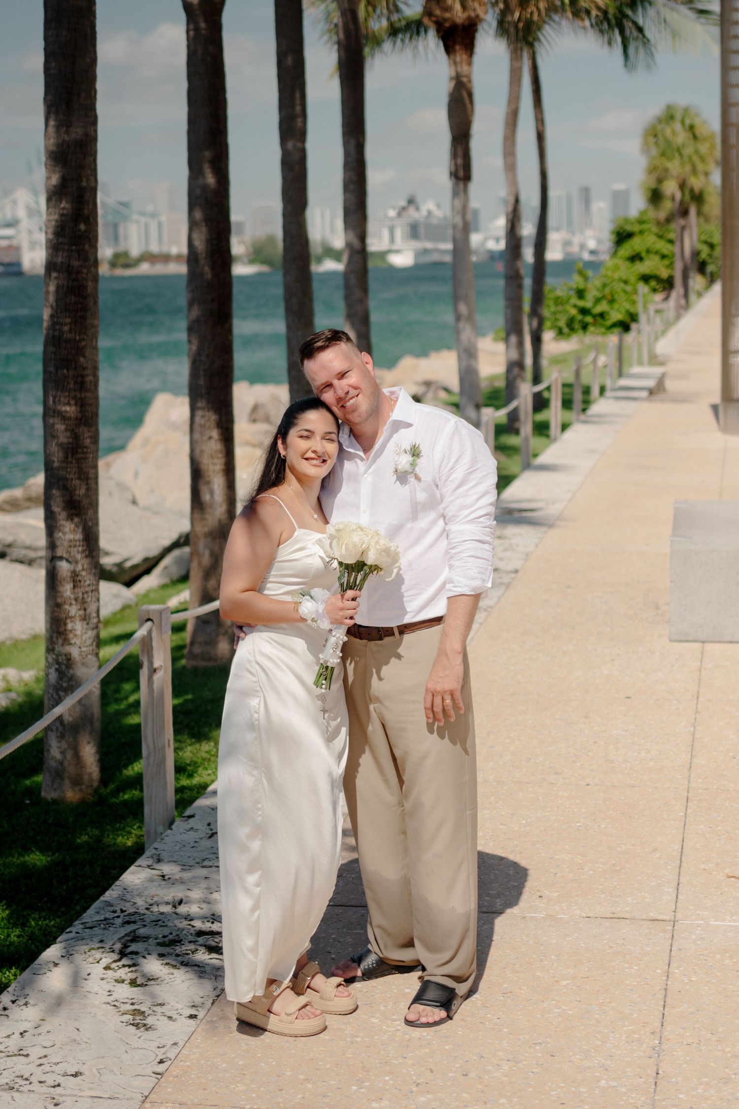 A couple in wedding attire poses playfully along a palm-lined beachfront walkway with ocean views in the background.