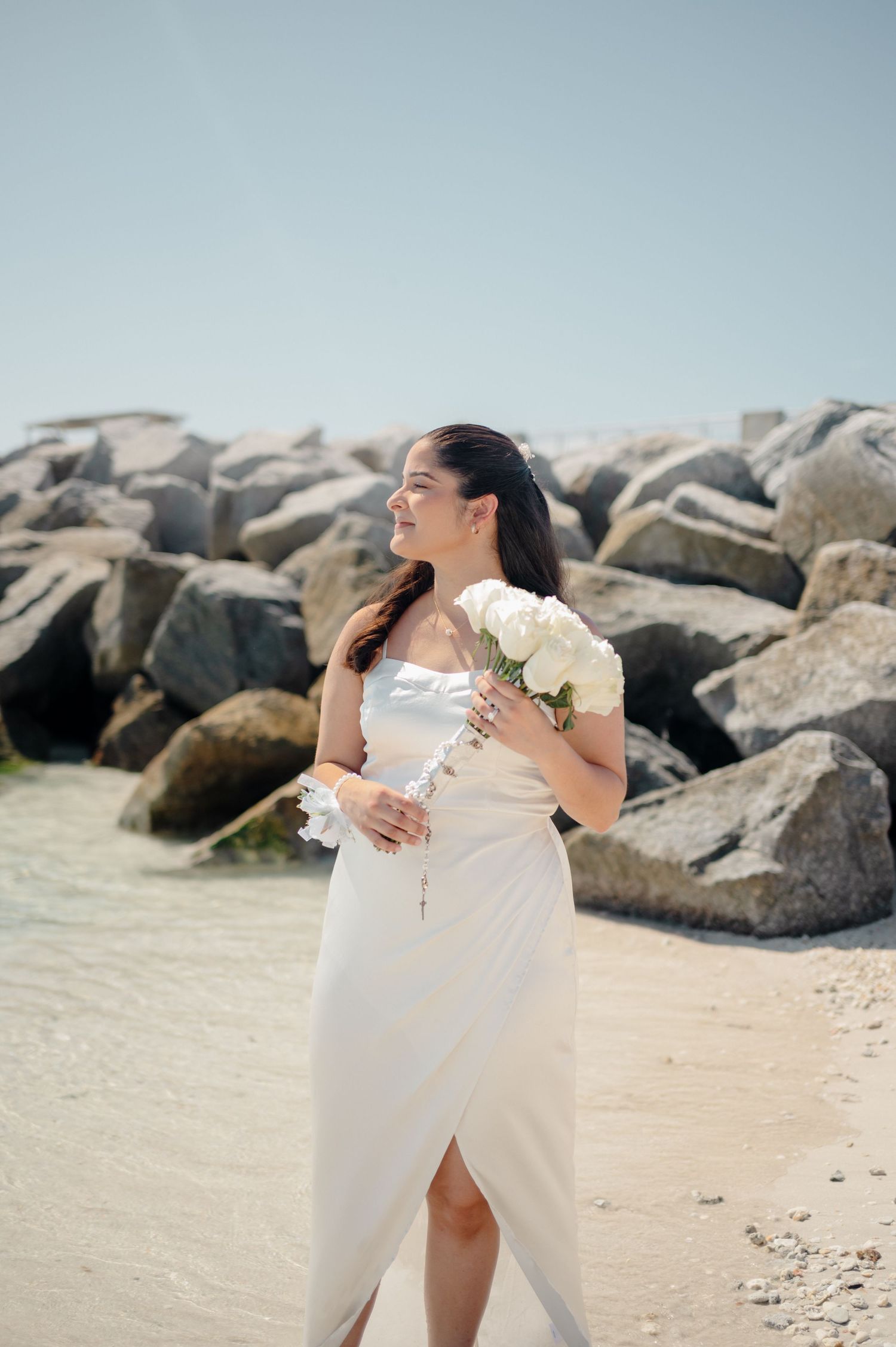 A couple in white wedding attire embraces and laughs together on a rocky beach during their coastal celebration.