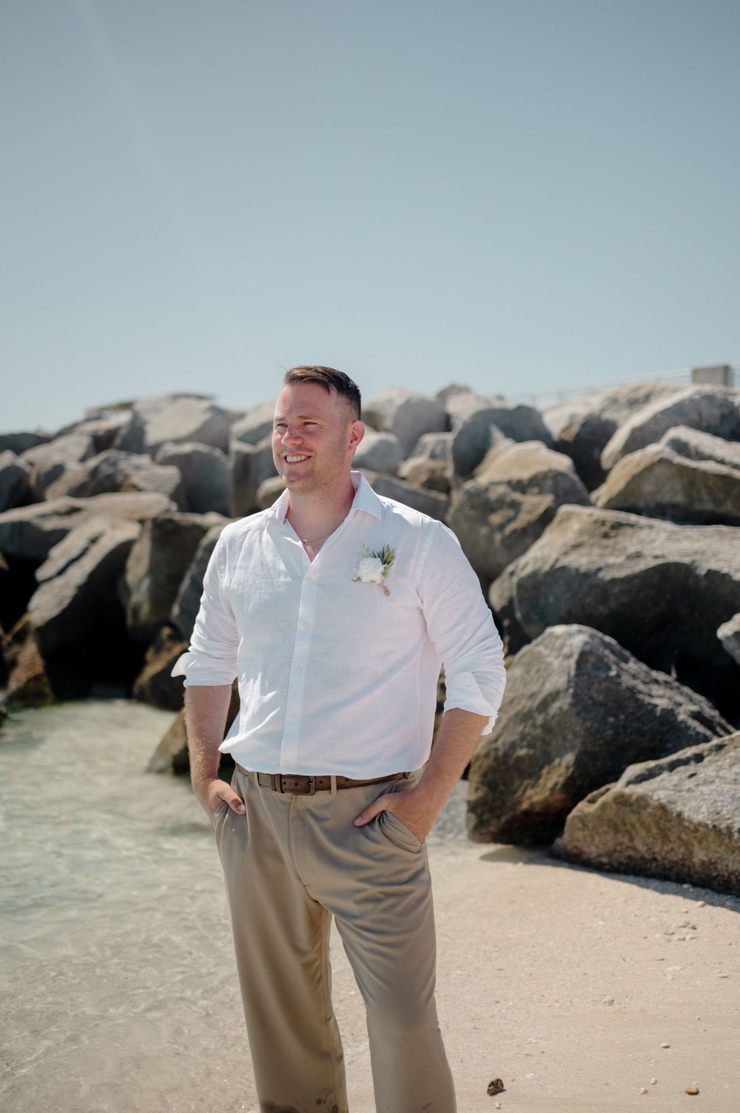 Person in white polo shirt and khaki pants poses on sandy beach with large rocky breakwater in background.