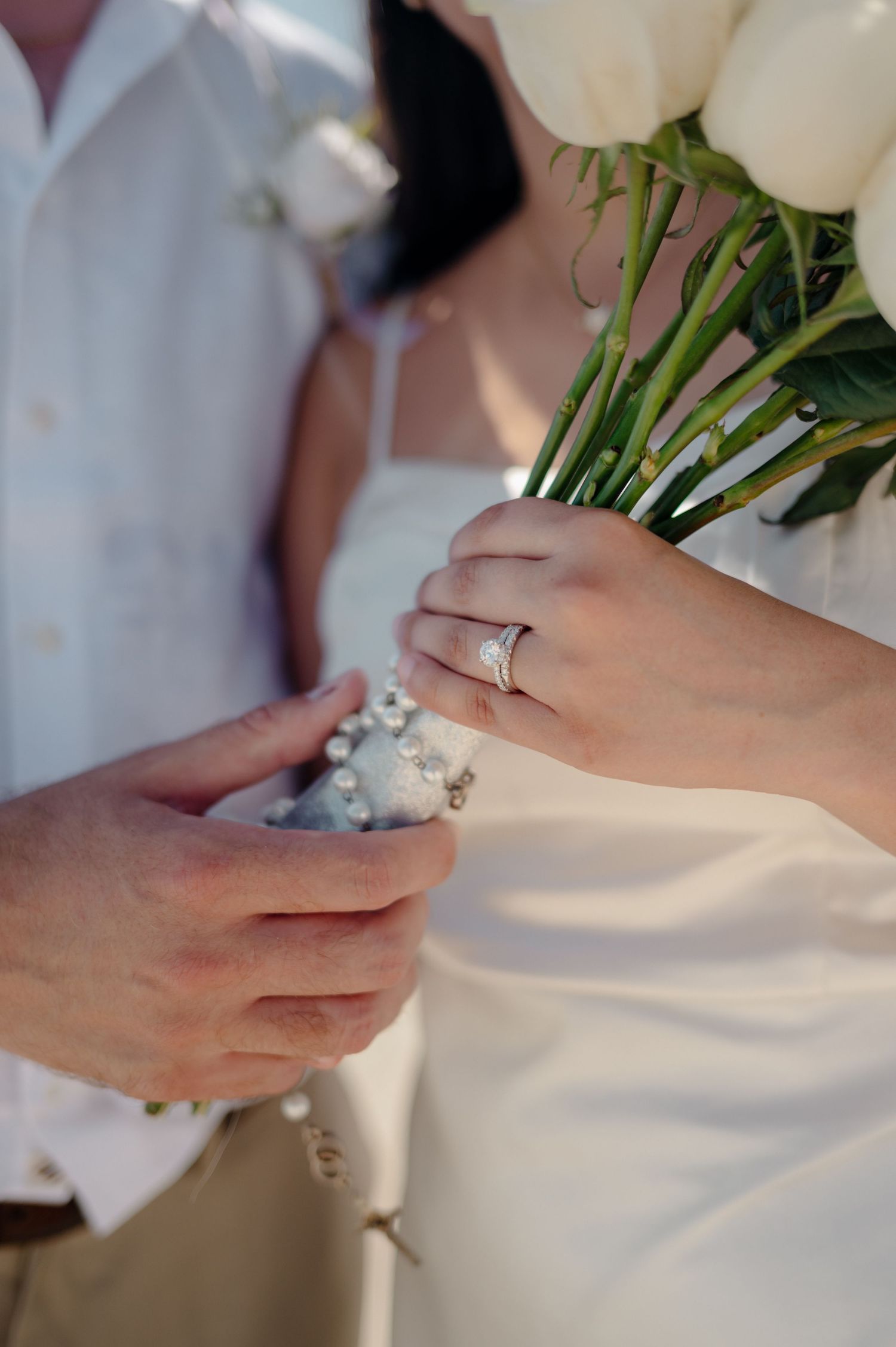 Close-up of hands exchanging wedding rings with white roses in bridal bouquet during ceremony.