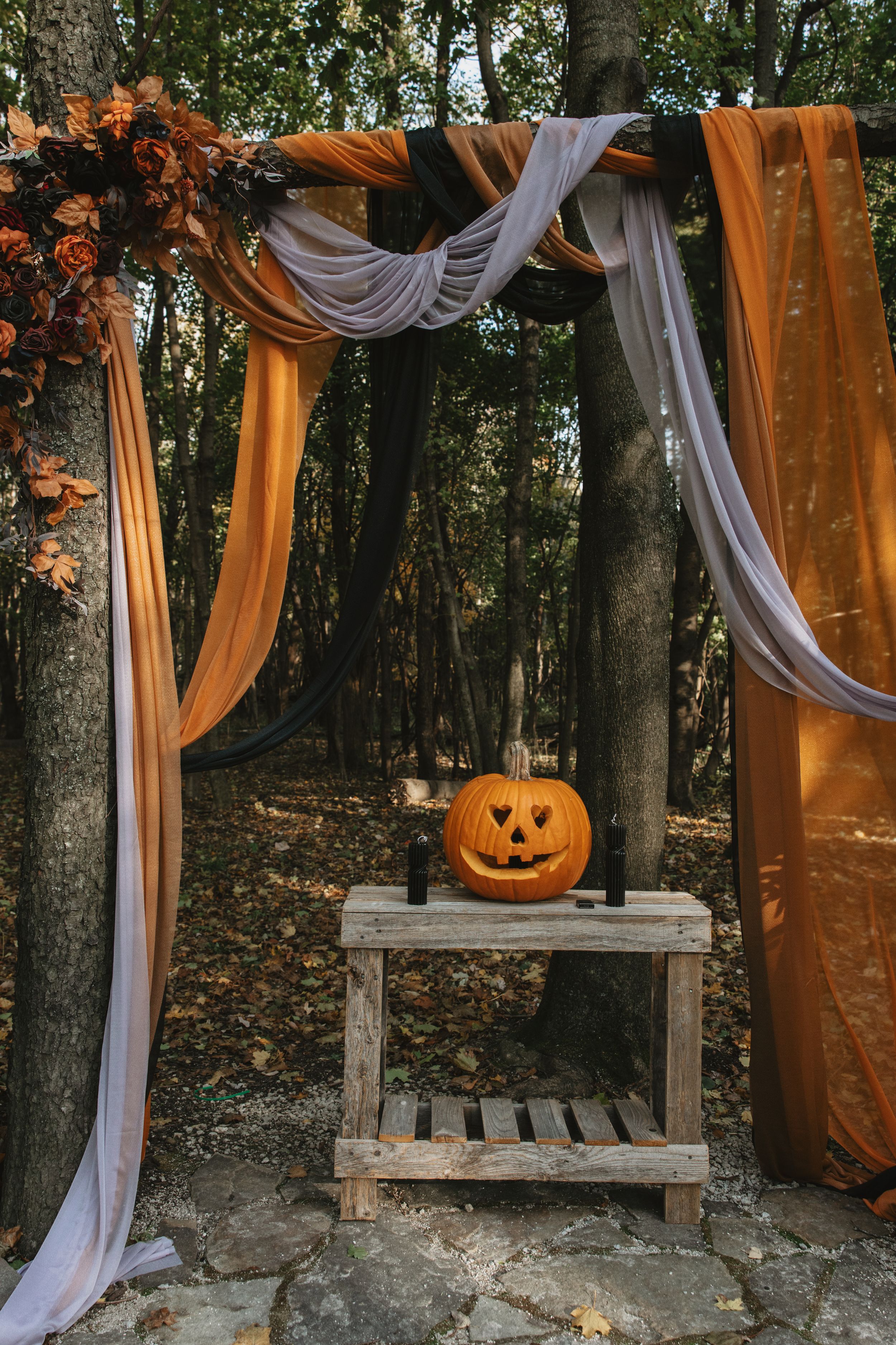 A carved pumpkin sits on a rustic wooden stand beneath an autumn-themed arch with orange and white fabric draping.