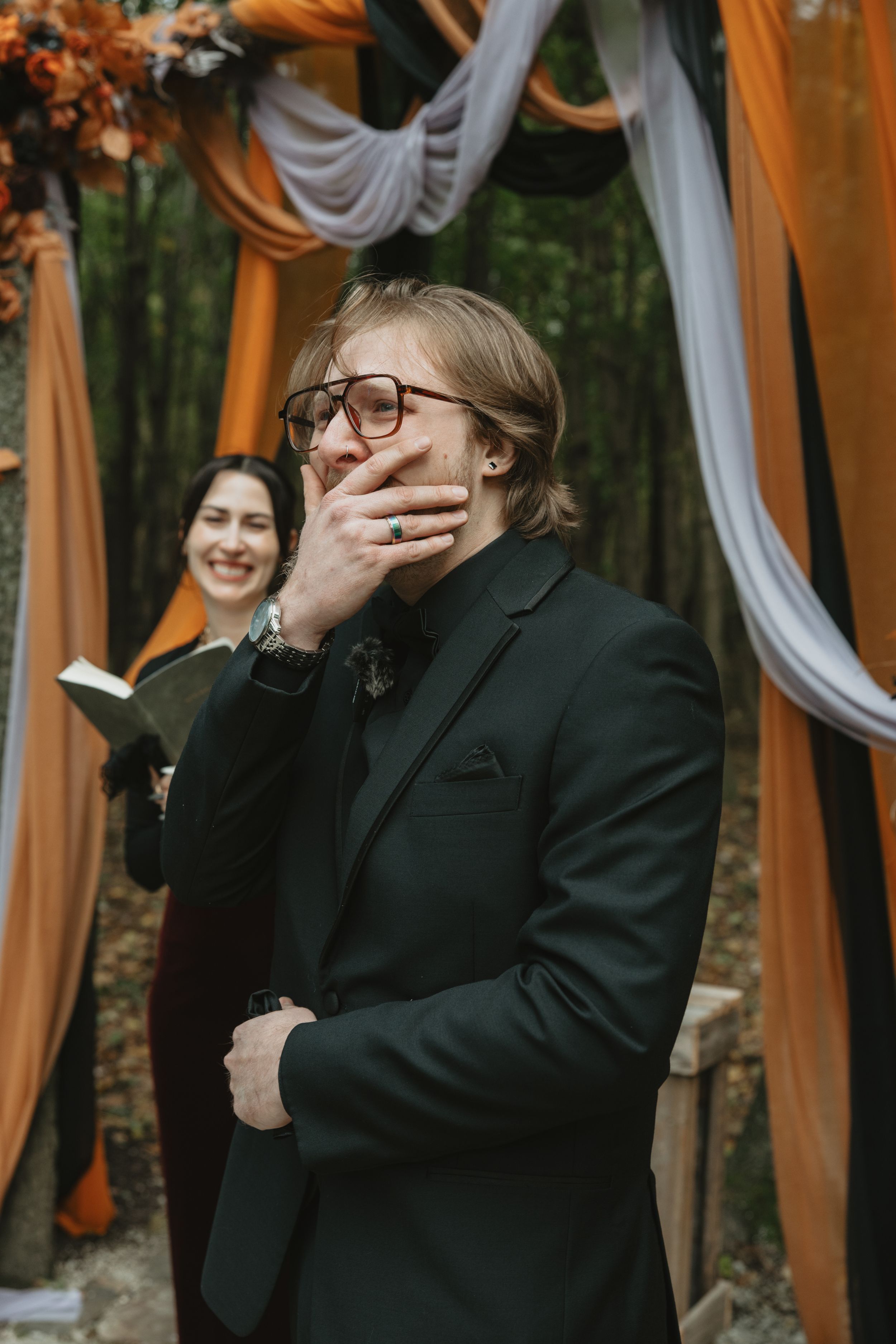 Emotional wedding scene under orange and white draped arch with candid reactions in black formal attire.