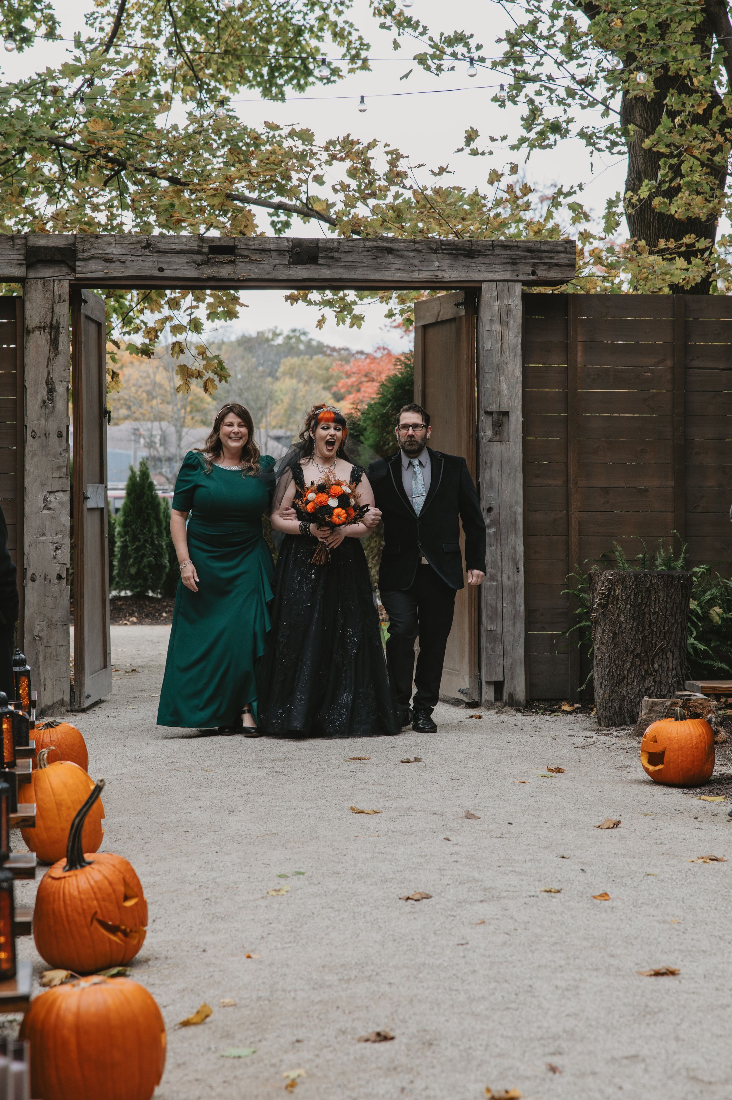 Wedding party poses along rustic wooden fence with autumn pumpkin decorations and fall foliage in background.
