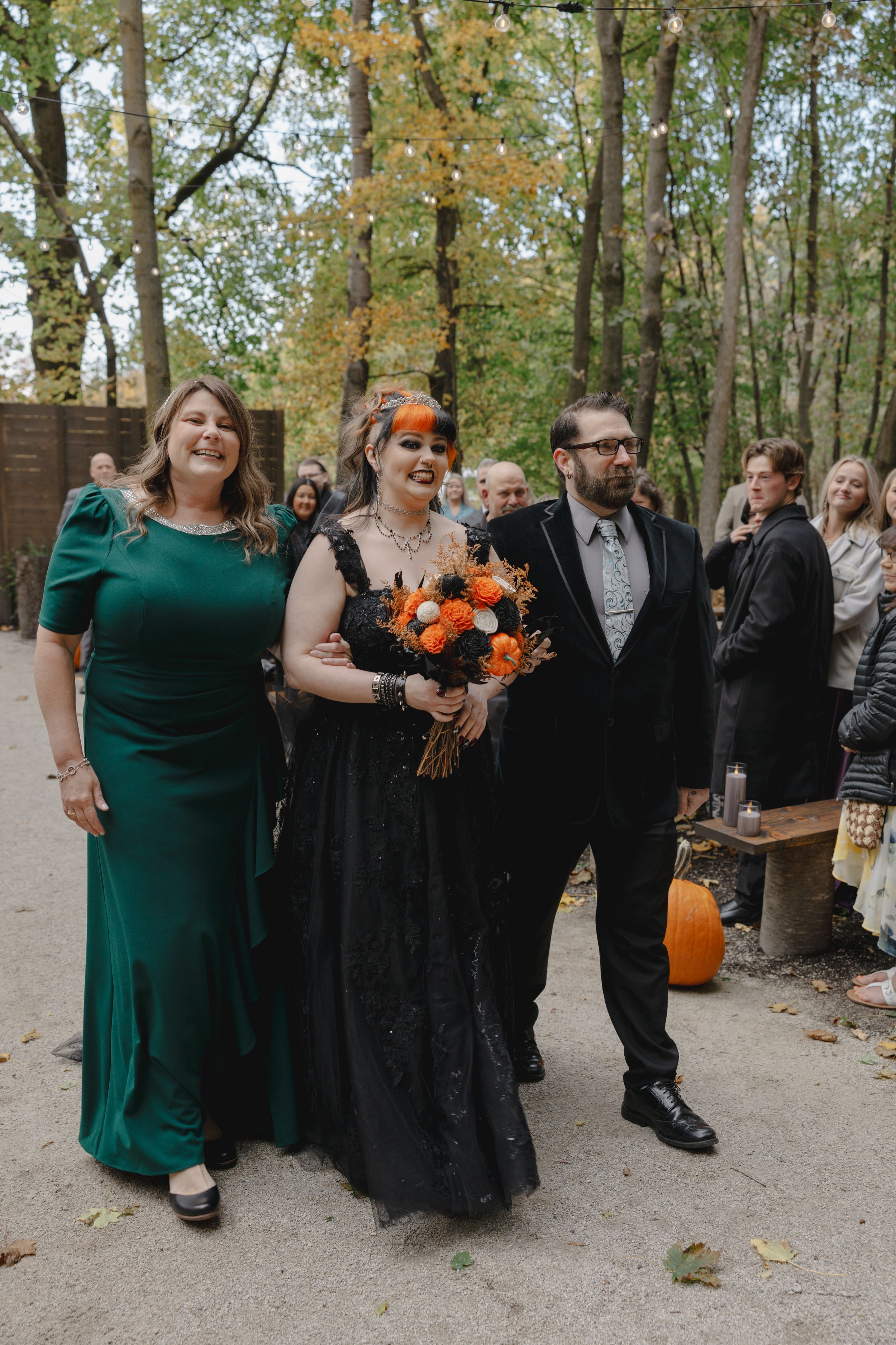Wedding party in emerald green dresses and black suits poses outdoors among autumn trees.