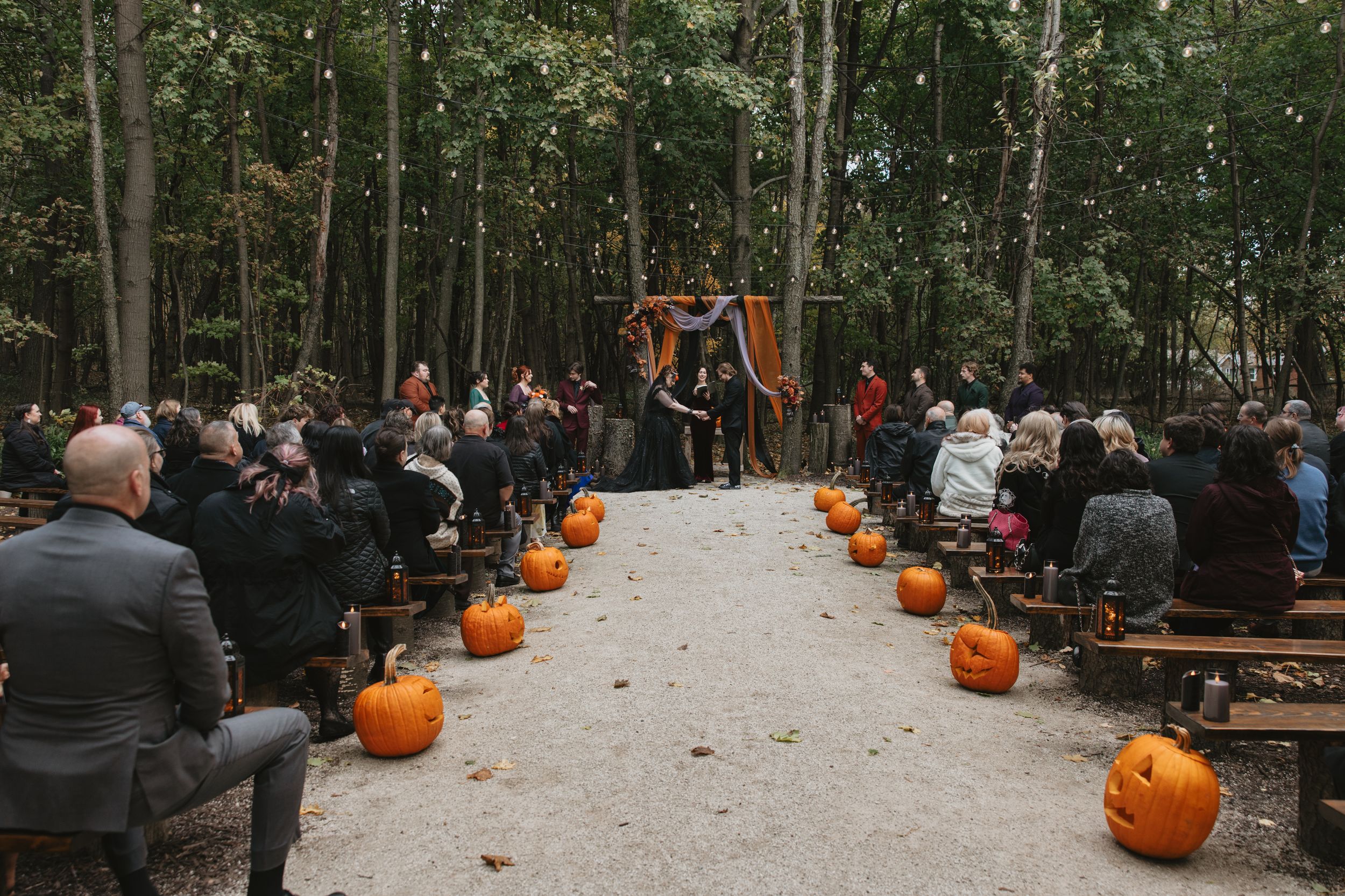 Outdoor fall wedding ceremony in a forest with pumpkins lining the aisle and guests seated on rustic wooden benches.