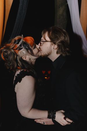 A romantic couple shares an intimate kiss in a dark moody setting with black curtains and gothic vibes.
