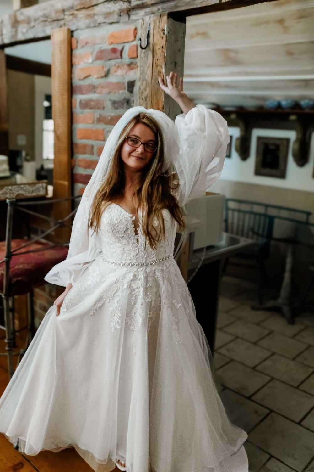 A bride in a white lace dress twirls playfully in an indoor rustic setting with exposed brick walls.
