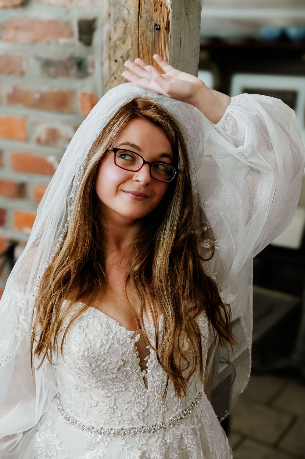 A bride in lace dress and veil poses playfully against rustic brick wall interior.