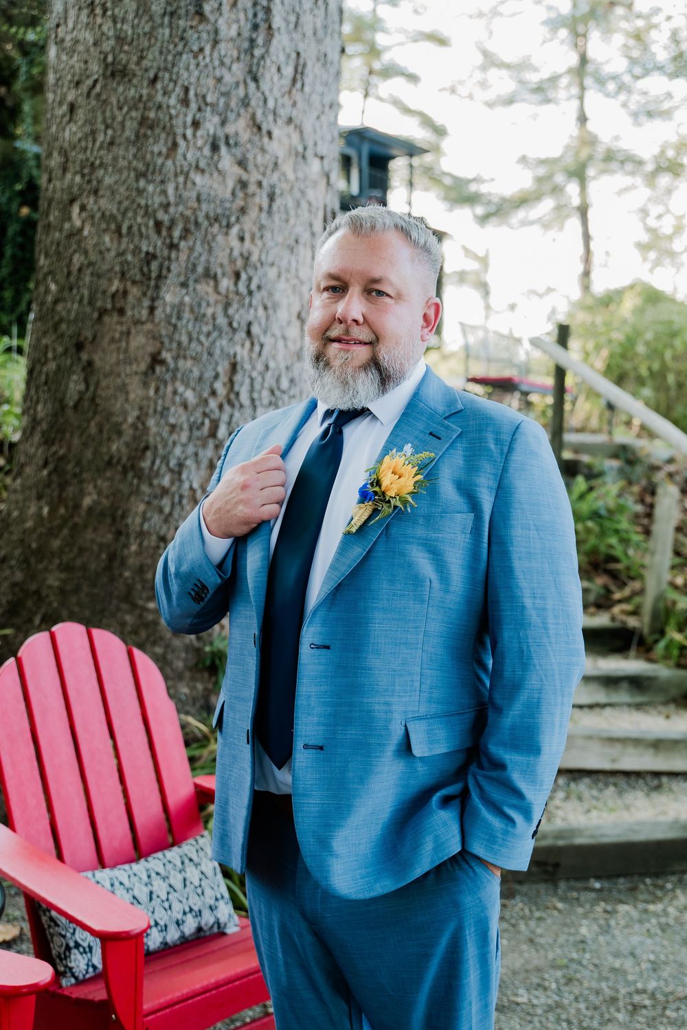 A groom in a light blue suit with yellow boutonniere poses near a red chair outdoors.