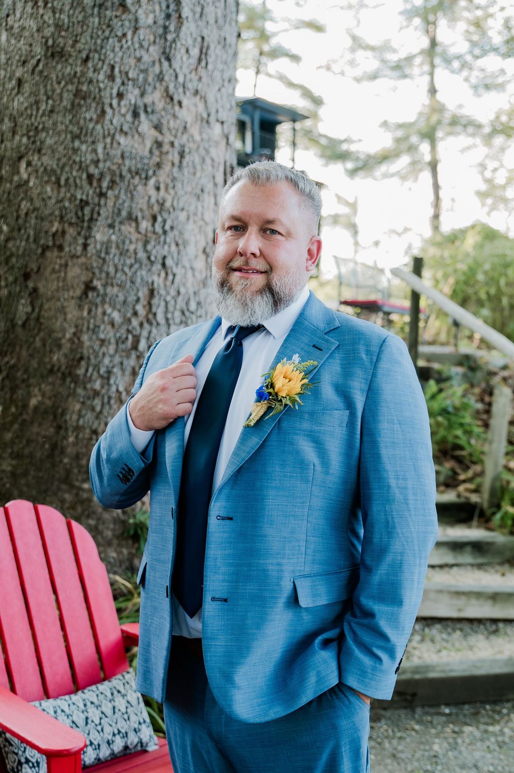 A groom in a bright blue suit stands confidently outdoors near a red Adirondack chair.