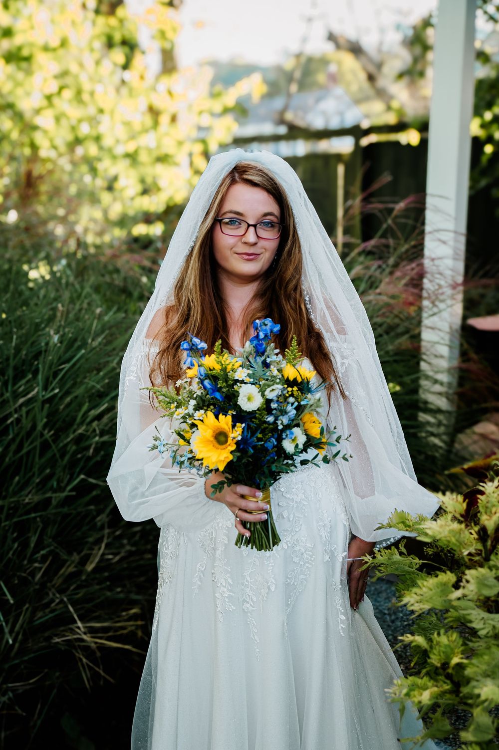 A bride in glasses holds a vibrant sunflower and blue flower bouquet in an outdoor garden setting.
