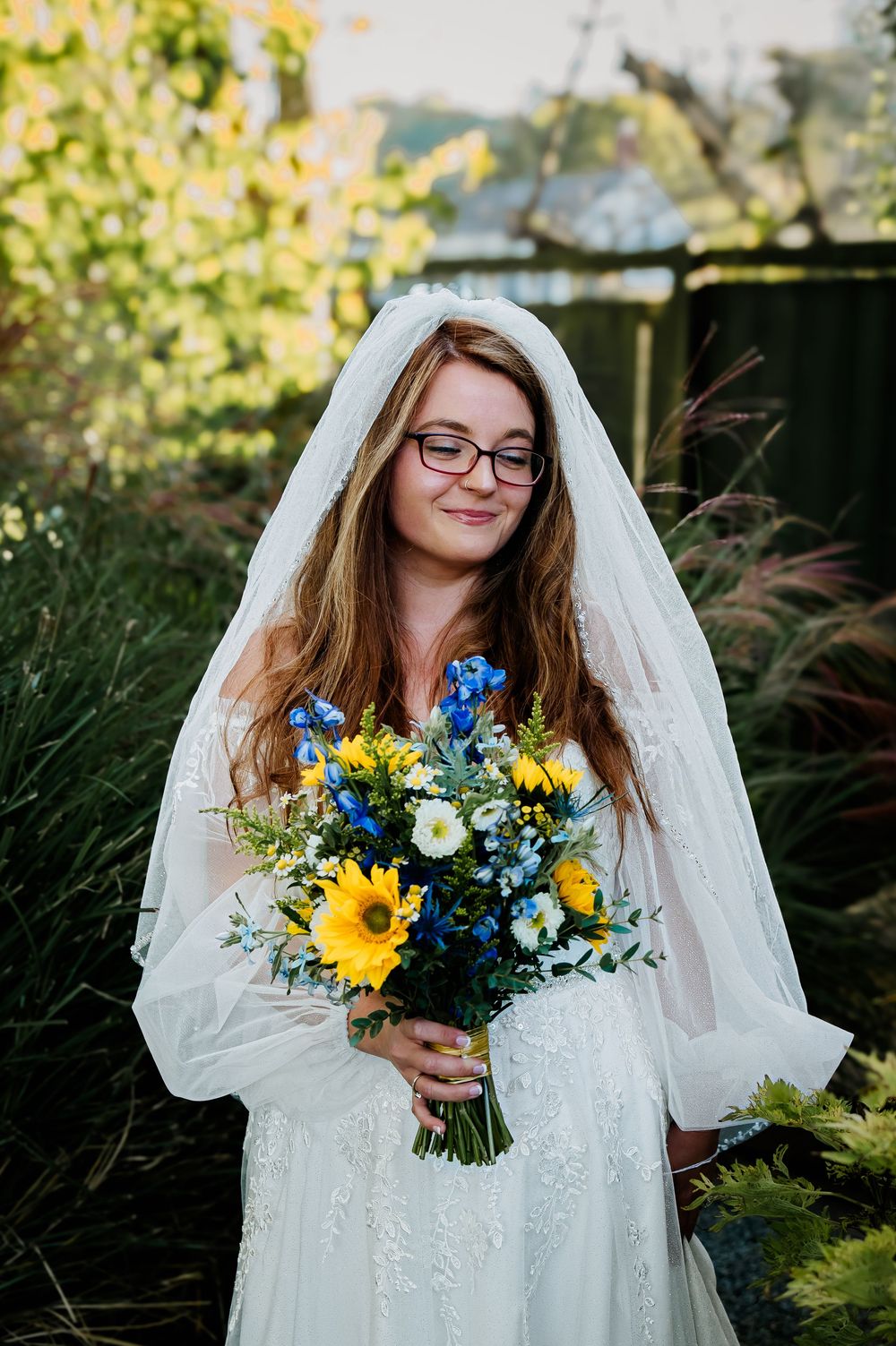 A bride wearing glasses smiles while holding a bouquet with sunflowers and blue flowers.