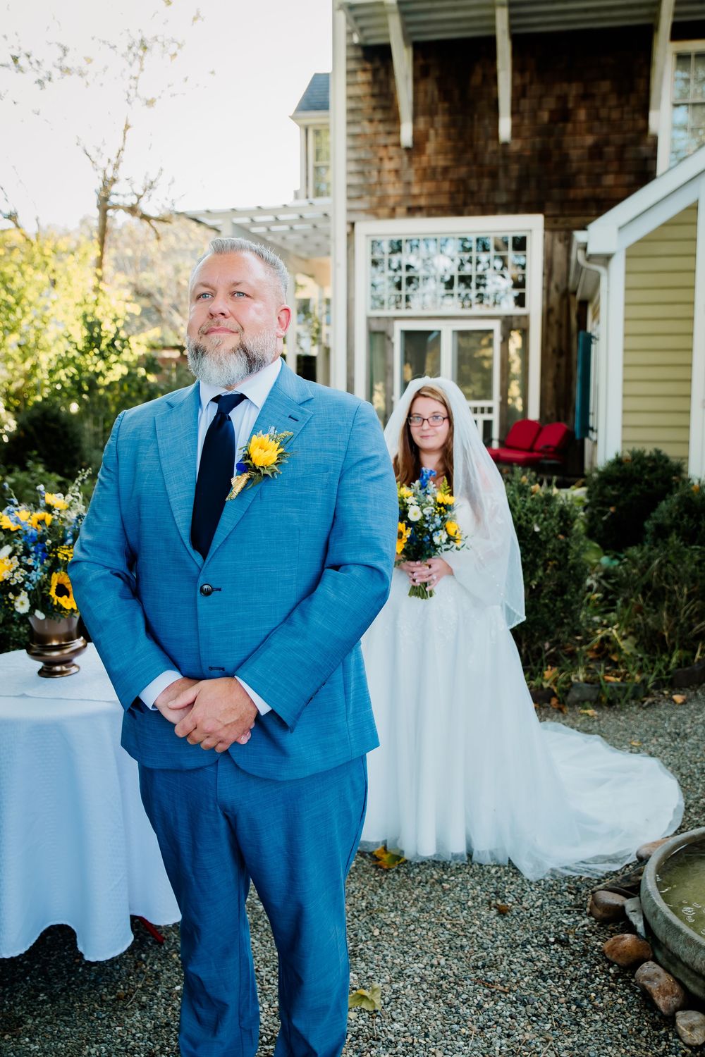 A wedding scene with a groom in blue suit and bride in white dress standing on gravel path.
