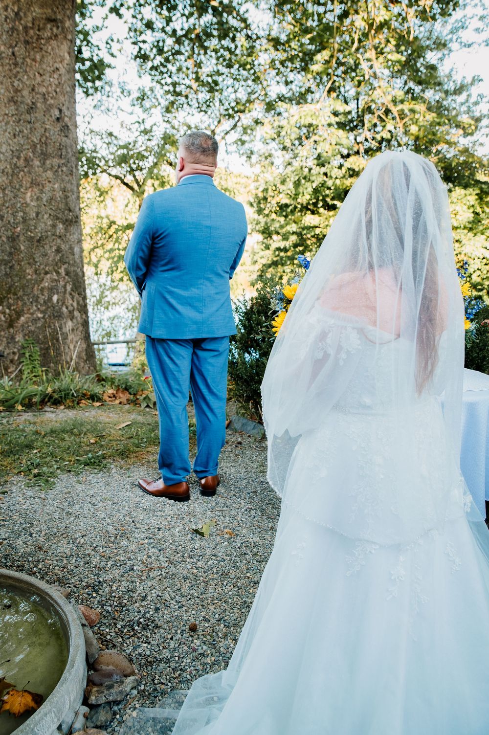 Wedding couple in blue suit and white dress photographed from behind in outdoor garden setting.
