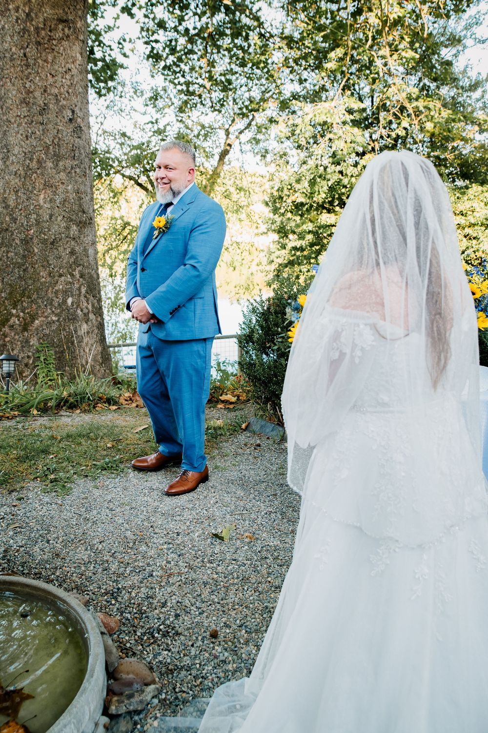 Emotional wedding first look scene with a blue-suited figure and white-veiled bride in an outdoor garden setting.