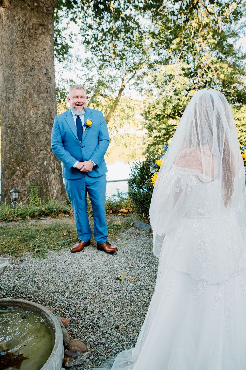 Wedding first look captured in a garden with sunlit trees, featuring a blue suit and flowing white bridal gown.