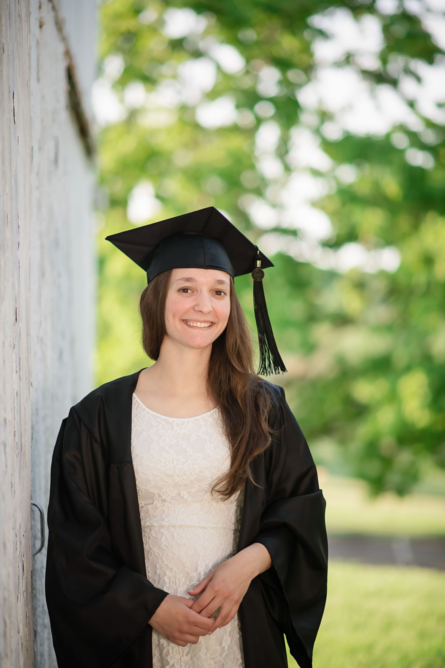 Graduate in cap and gown poses against white wall with green foliage background.