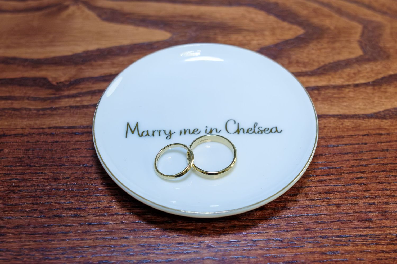 Gold wedding rings rest on a white ceramic dish with handwritten text against a rich wooden surface.