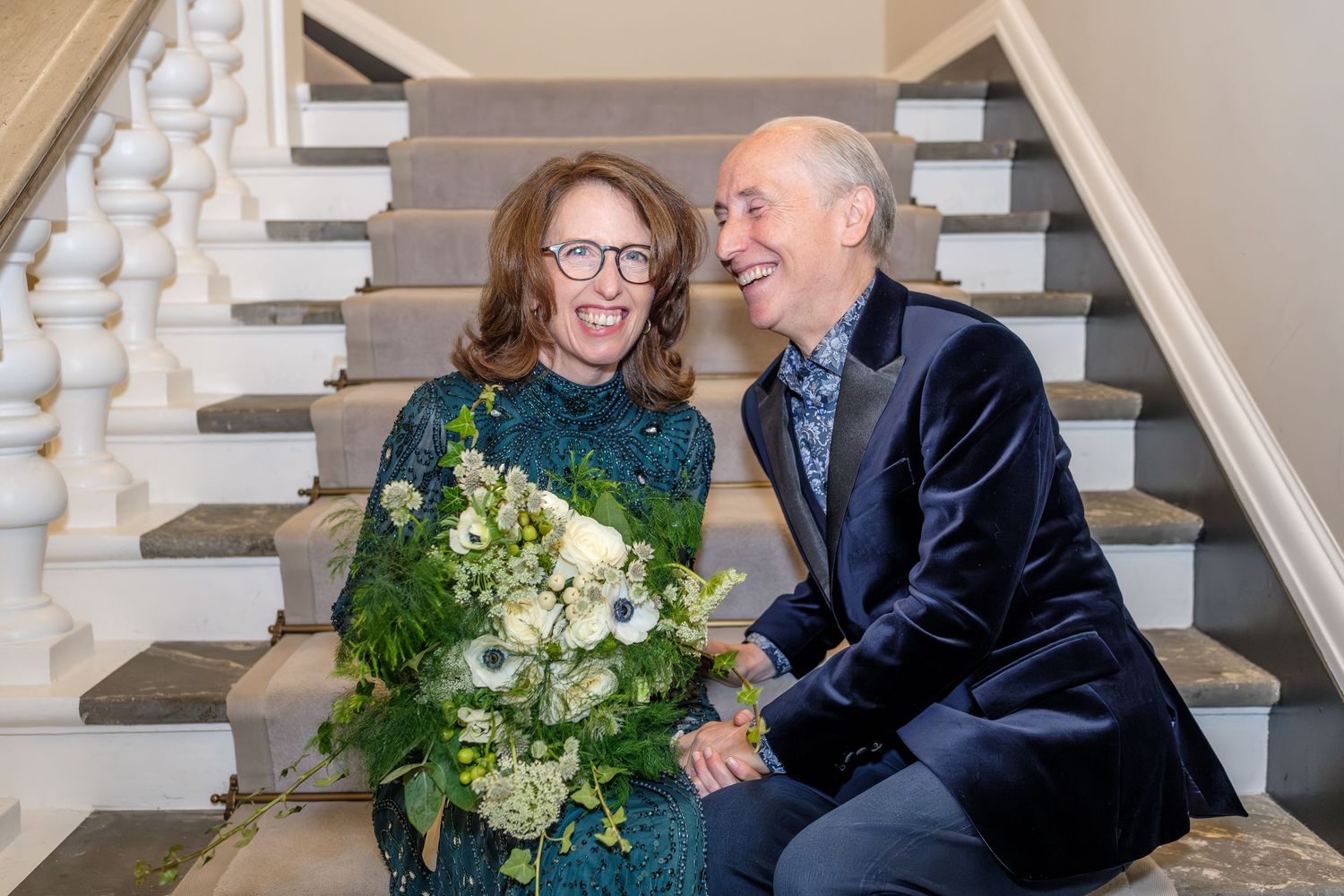 A couple shares a tender moment on a staircase with white railings, the woman holding a bouquet of white flowers.