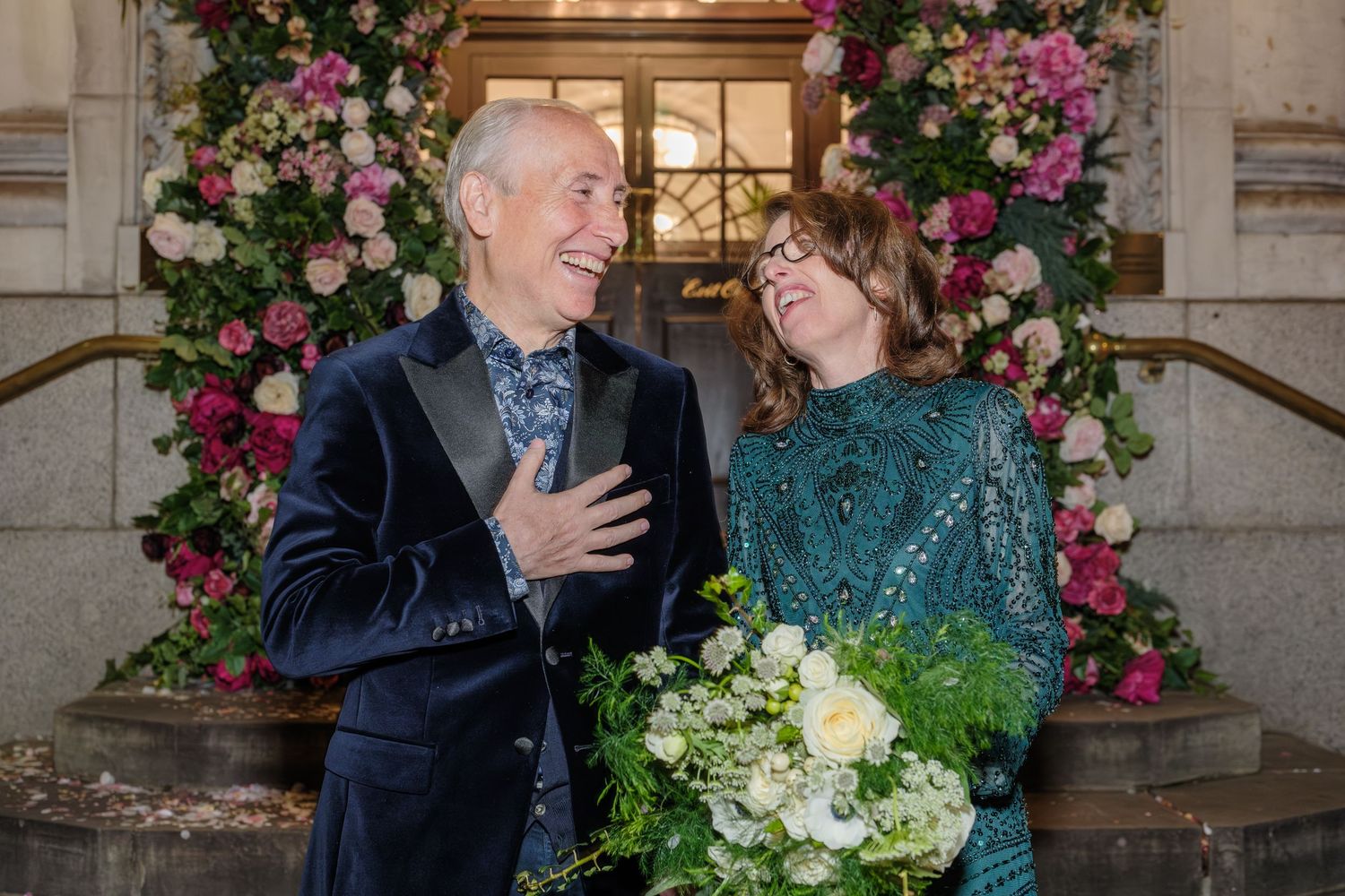 Two people share a joyful moment in front of an elegant floral arch with pink roses and white bouquet.