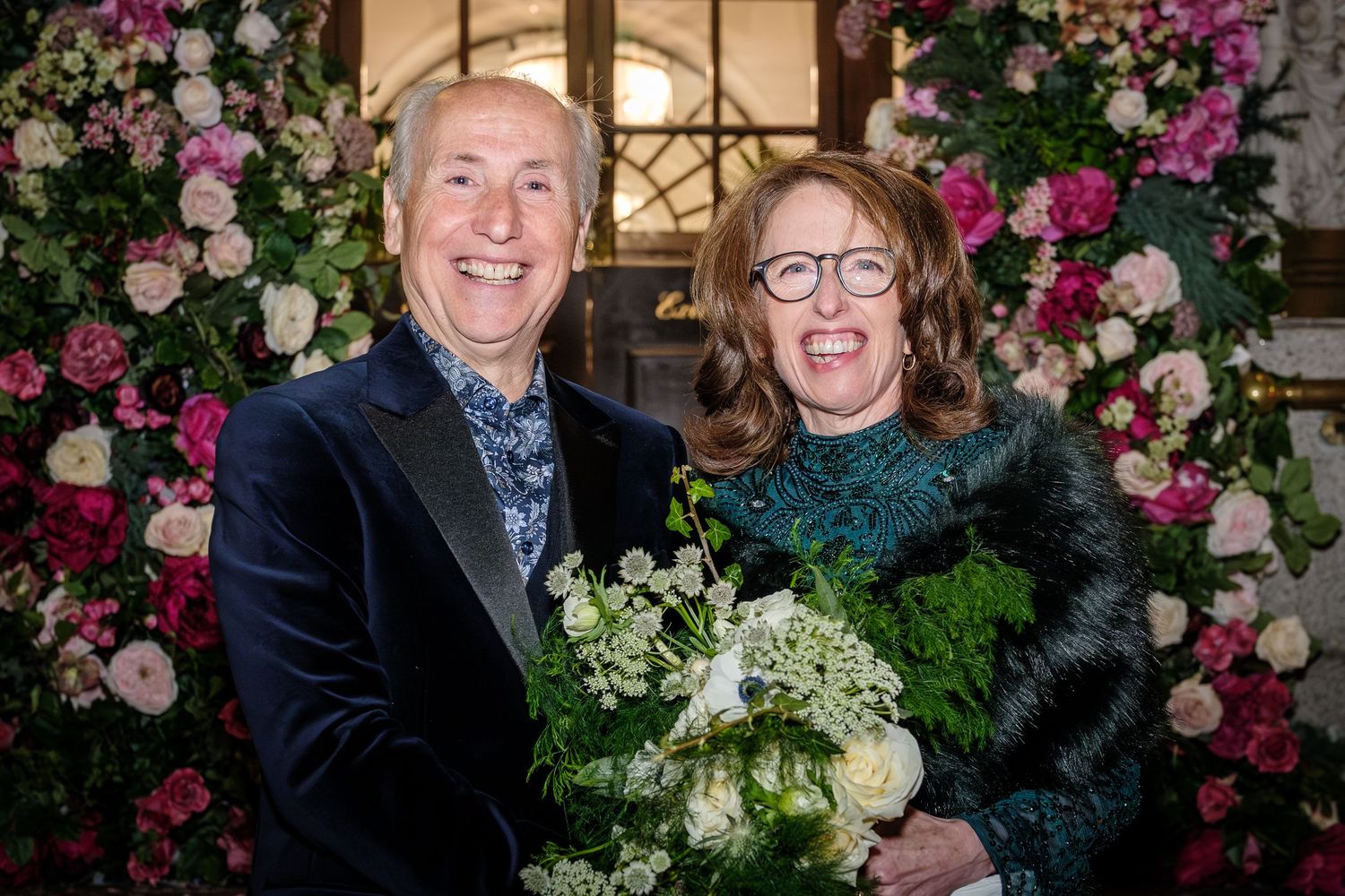 A couple poses together at a floral-decorated event venue with pink and white roses in the background.