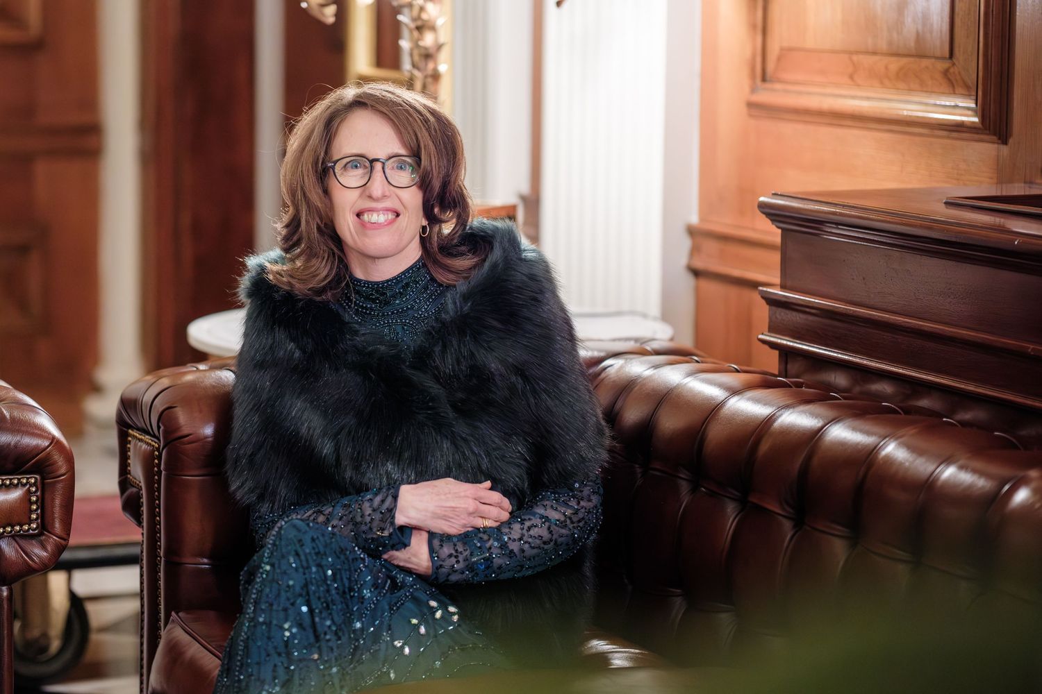 A smiling person wearing glasses sits in a brown leather chair at a formal wooden desk.