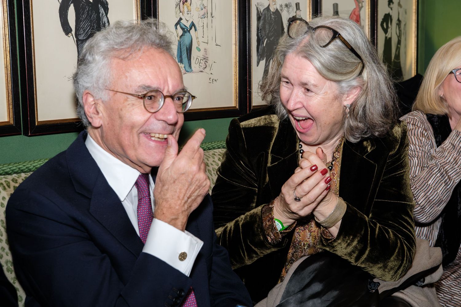 Two elegantly dressed seniors share a lighthearted moment while laughing together at a social gathering.