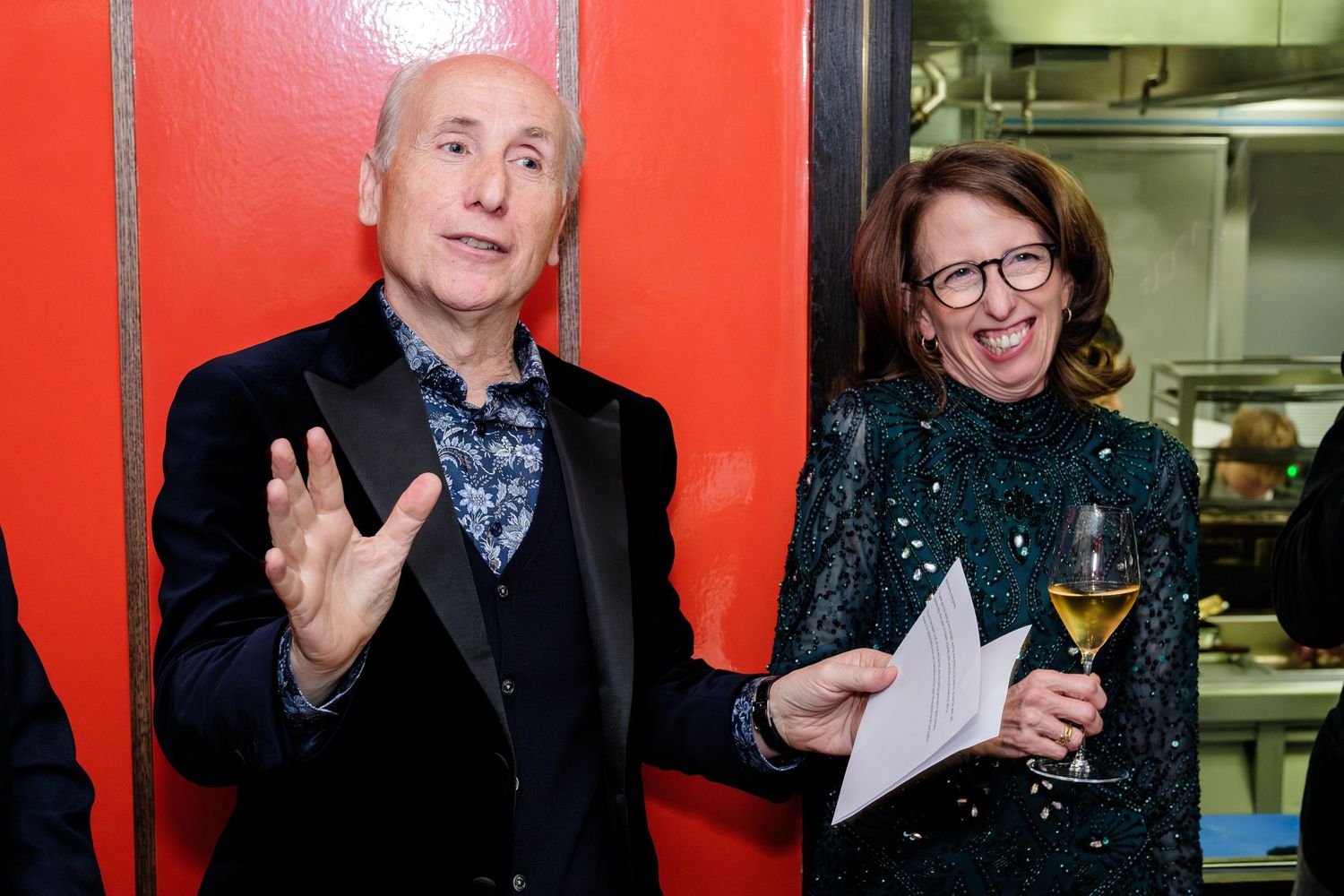 Two elegantly dressed people at an event, one in a black suit with bow tie gesturing and one in a sparkly green outfit.