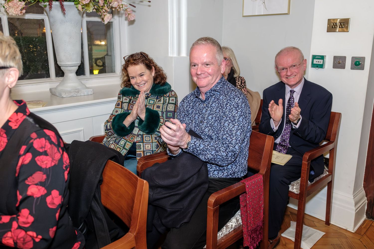 A group of people applauding while seated in wooden chairs at an indoor gathering.