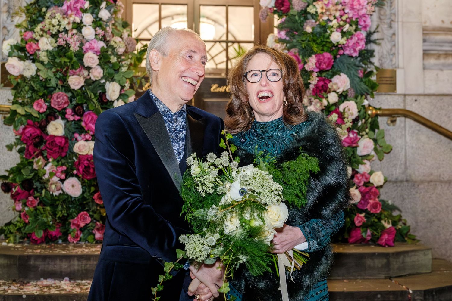 A couple shares a joyful moment together surrounded by pink roses and floral arrangements at an elegant venue.