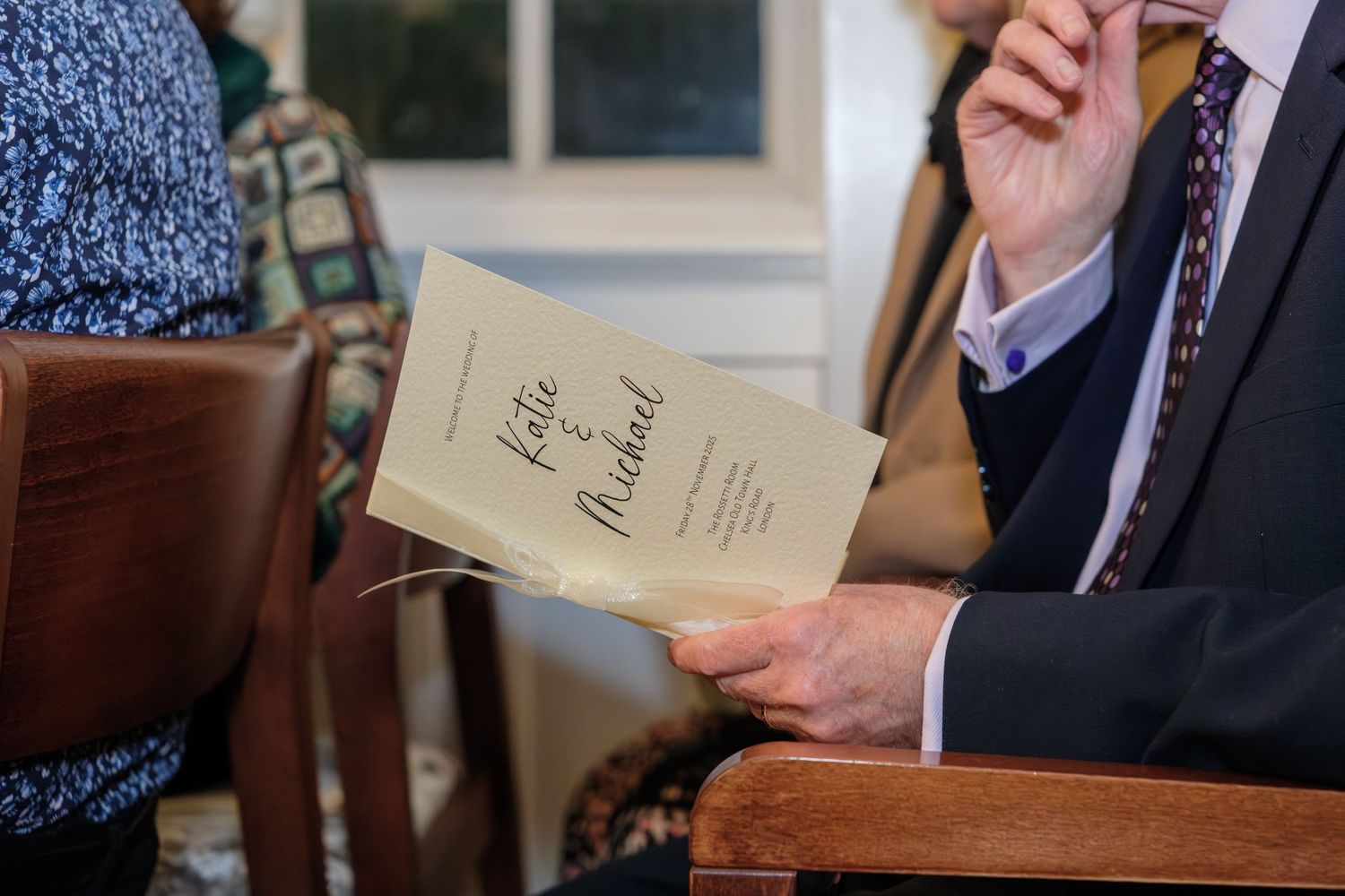 Someone in a suit holds a vintage notebook or document while seated in a wooden chair.