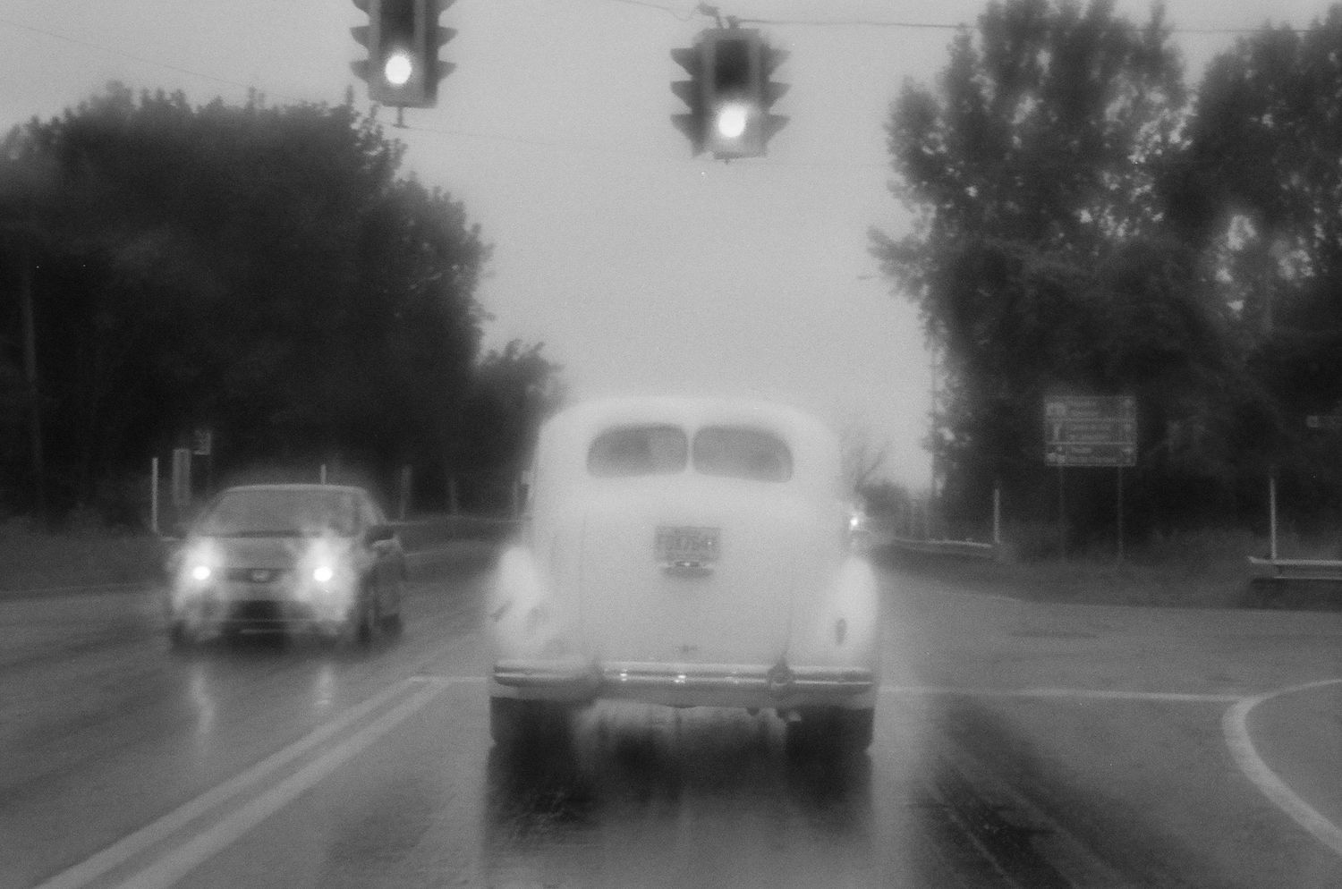 Vintage car drives on a misty road with traffic lights and trees visible in black and white photograph.