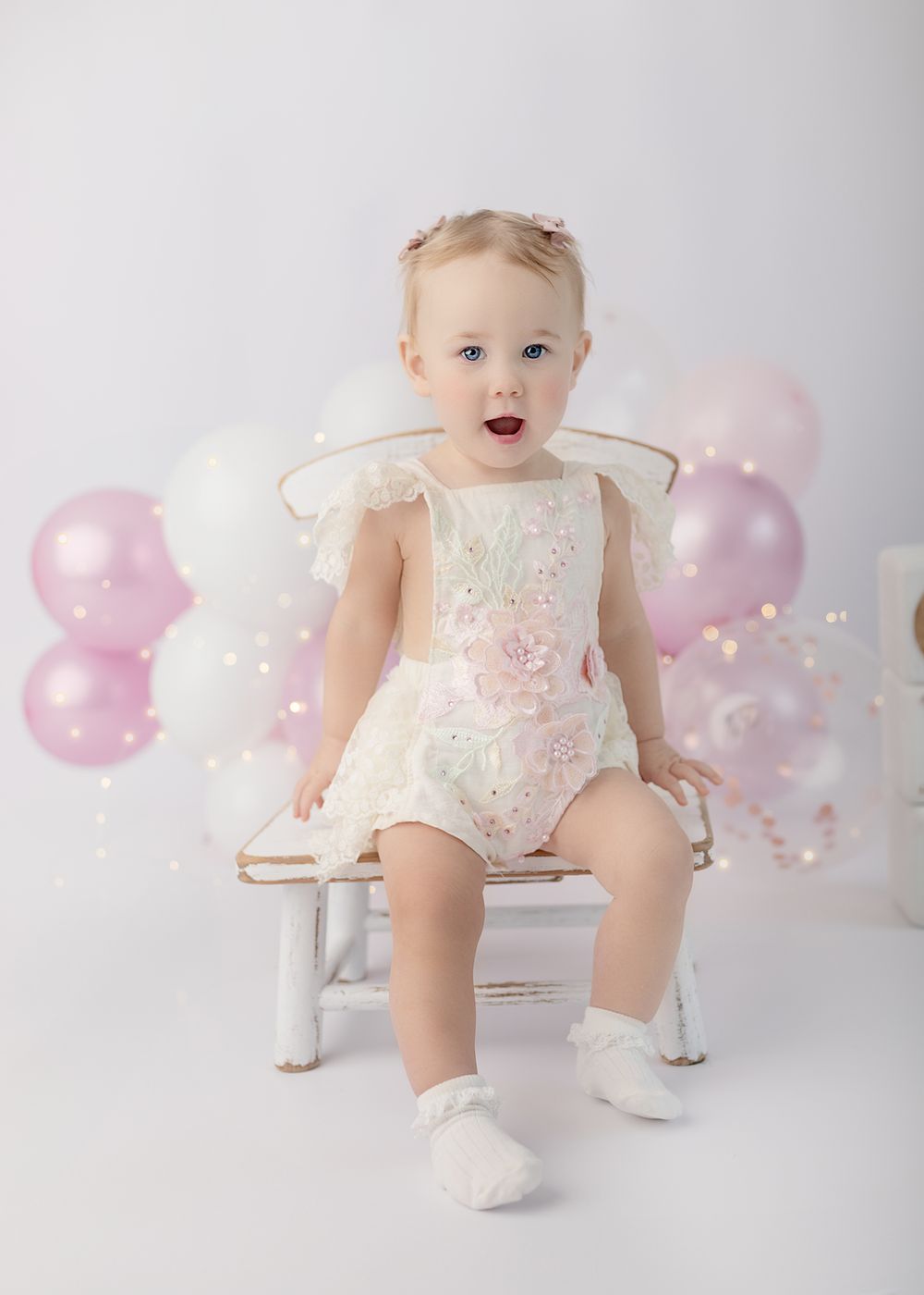 Baby in white lace outfit sits on vintage chair with pink and white balloons in dreamy studio setting.