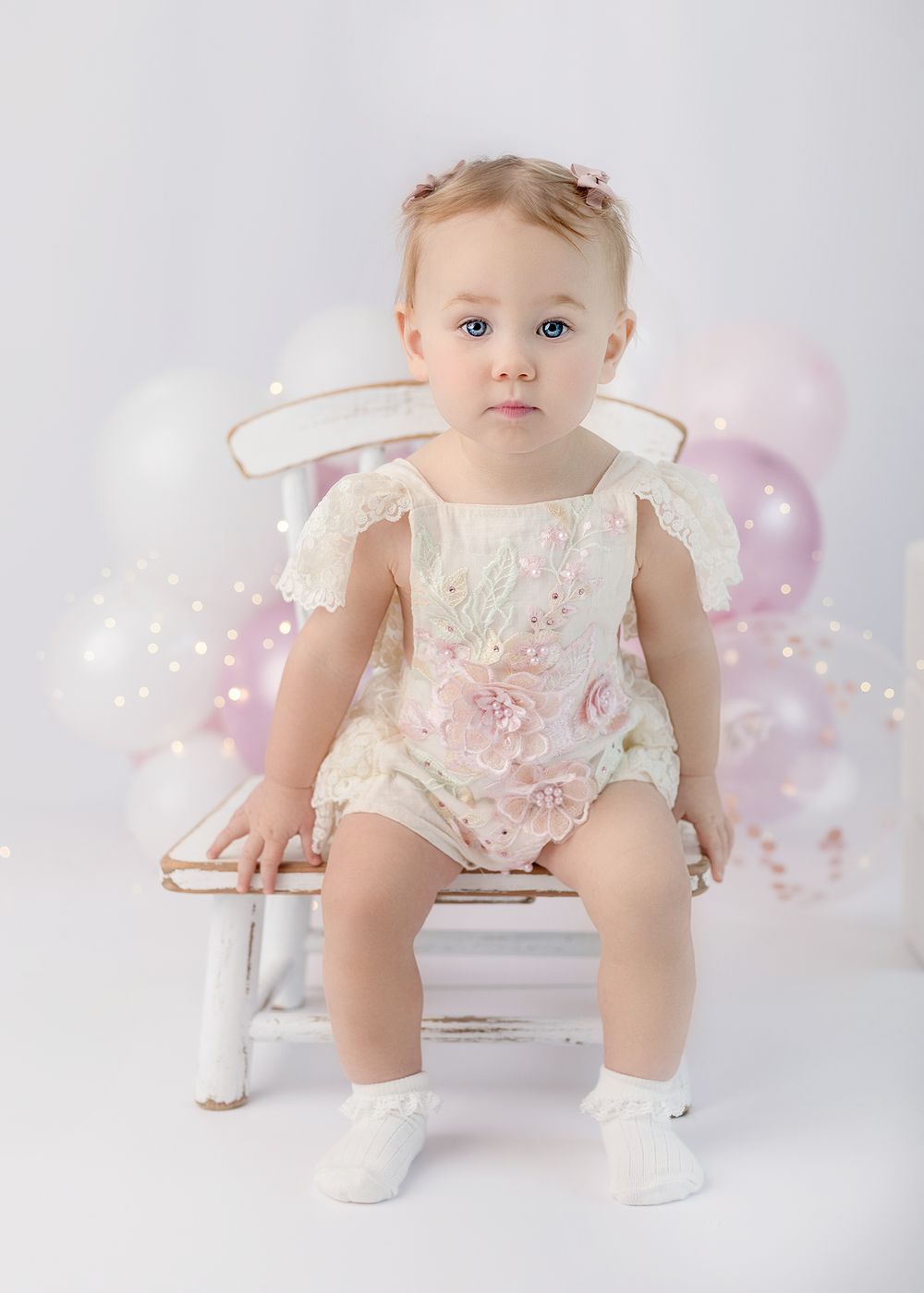 Baby wearing floral romper sits on white bench with pink balloons and decorative party elements in background.