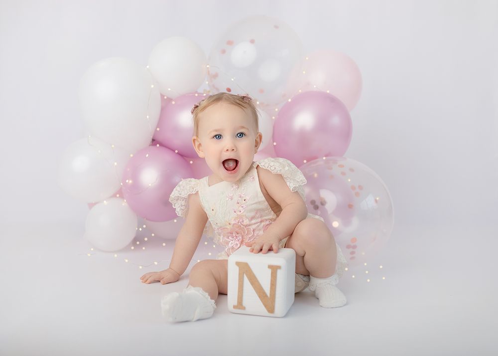 Baby in white outfit smiles joyfully surrounded by pink and white balloons during cake smash photo session.