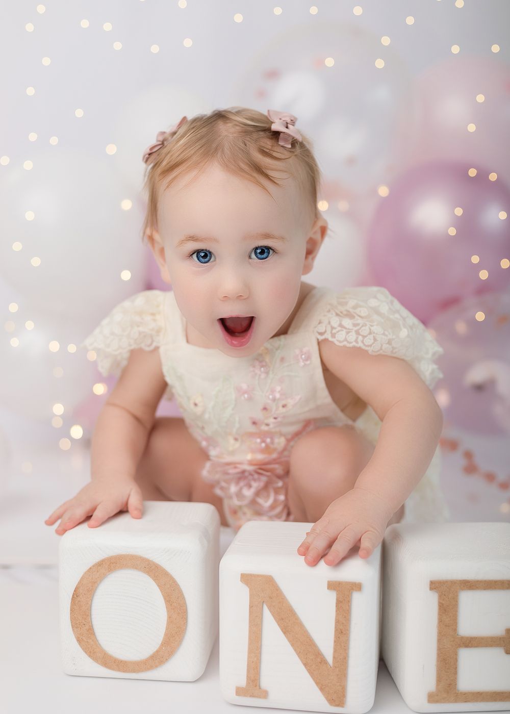 A baby in a white lace dress leans on decorative wooden blocks spelling 'ONE' against a sparkly bokeh background.