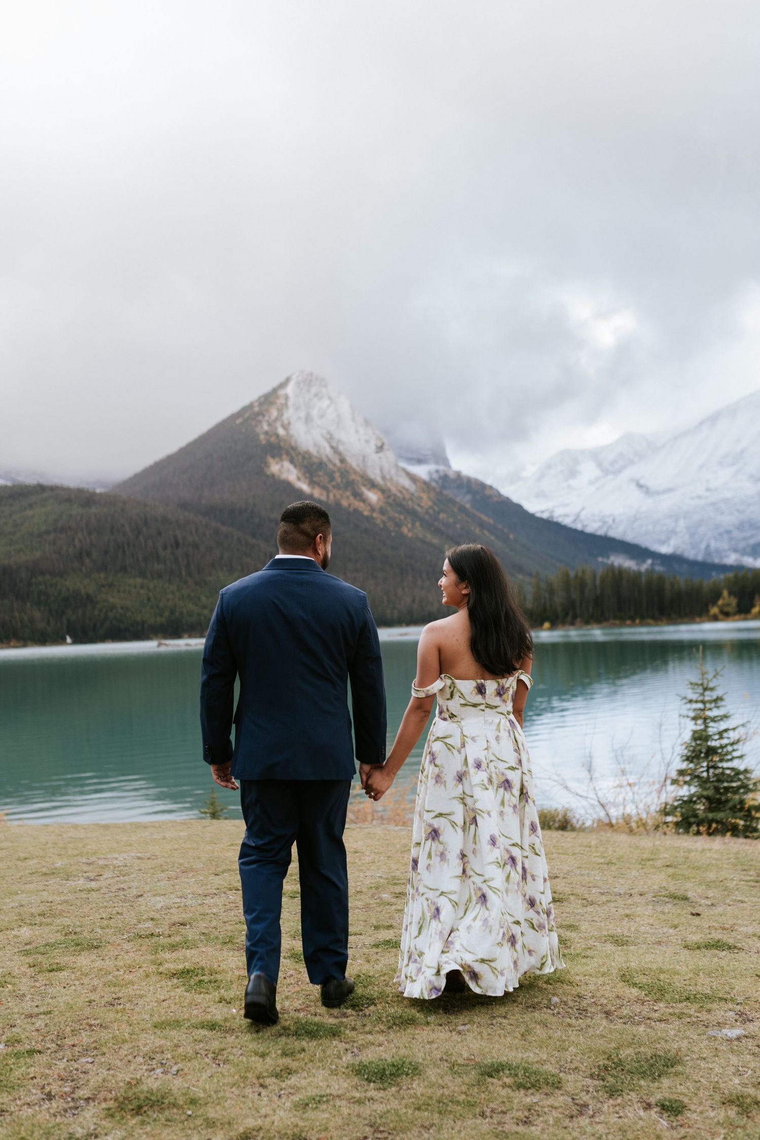 A couple in wedding attire stands together overlooking a scenic mountain lake landscape in multiple similar poses.