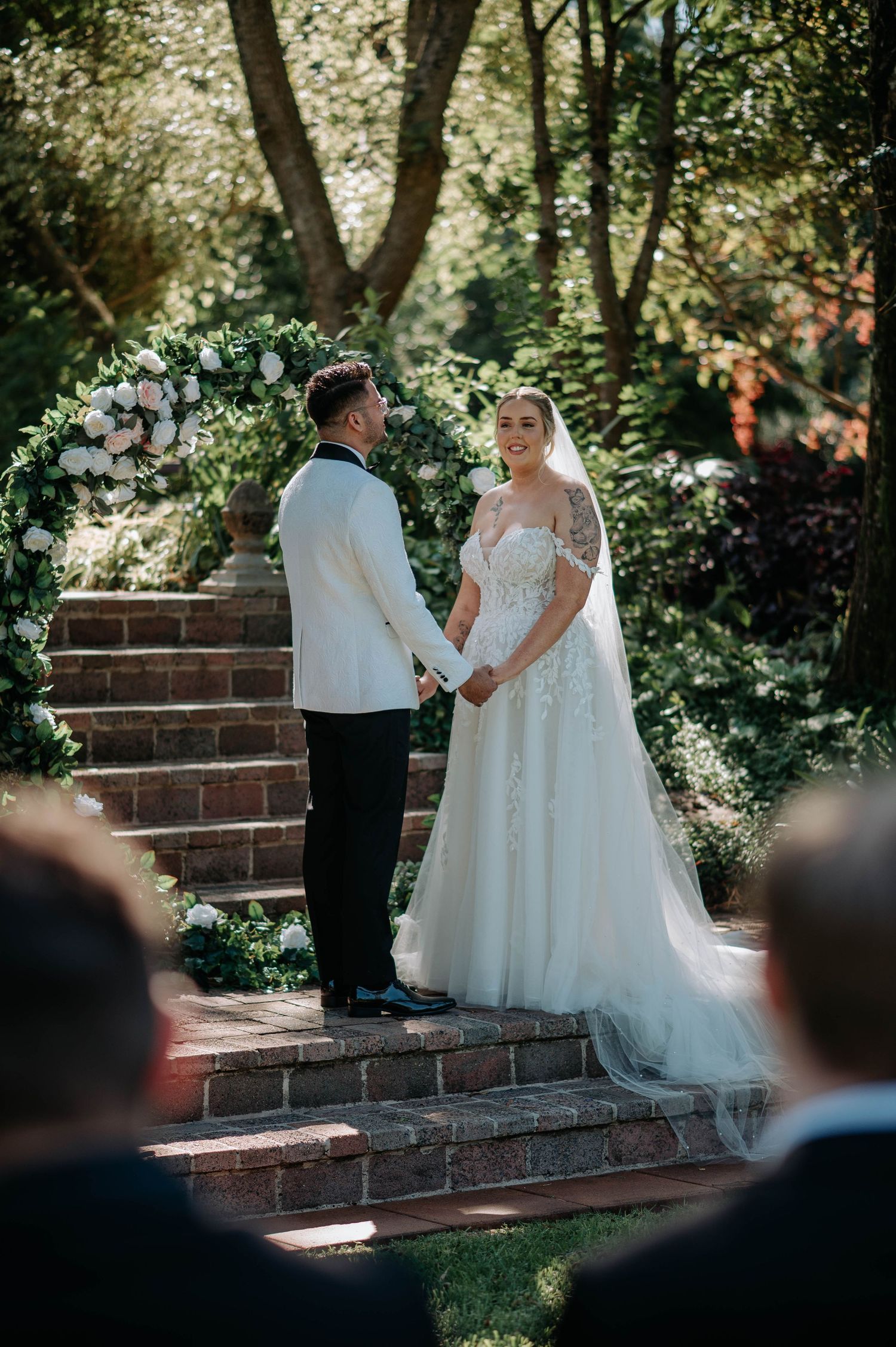 A wedding couple shares an intimate moment on brick steps surrounded by lush garden greenery in natural sunlight.
