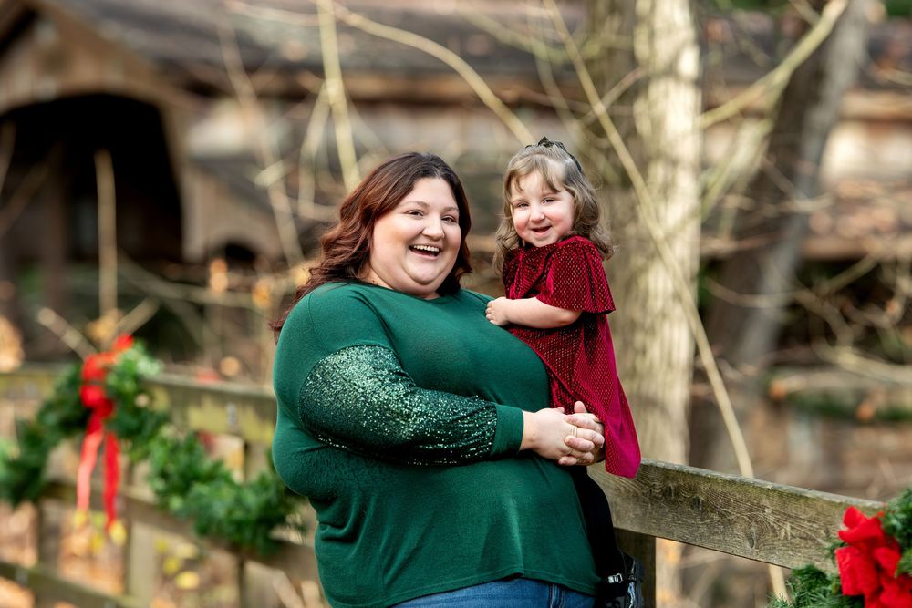 A smiling mother in a green top holds her young daughter in a red dress near Christmas wreaths outdoors.