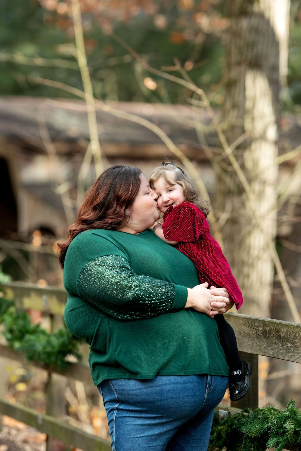 A mother lovingly kisses her young daughter in a red dress while standing near a wooden fence outdoors.