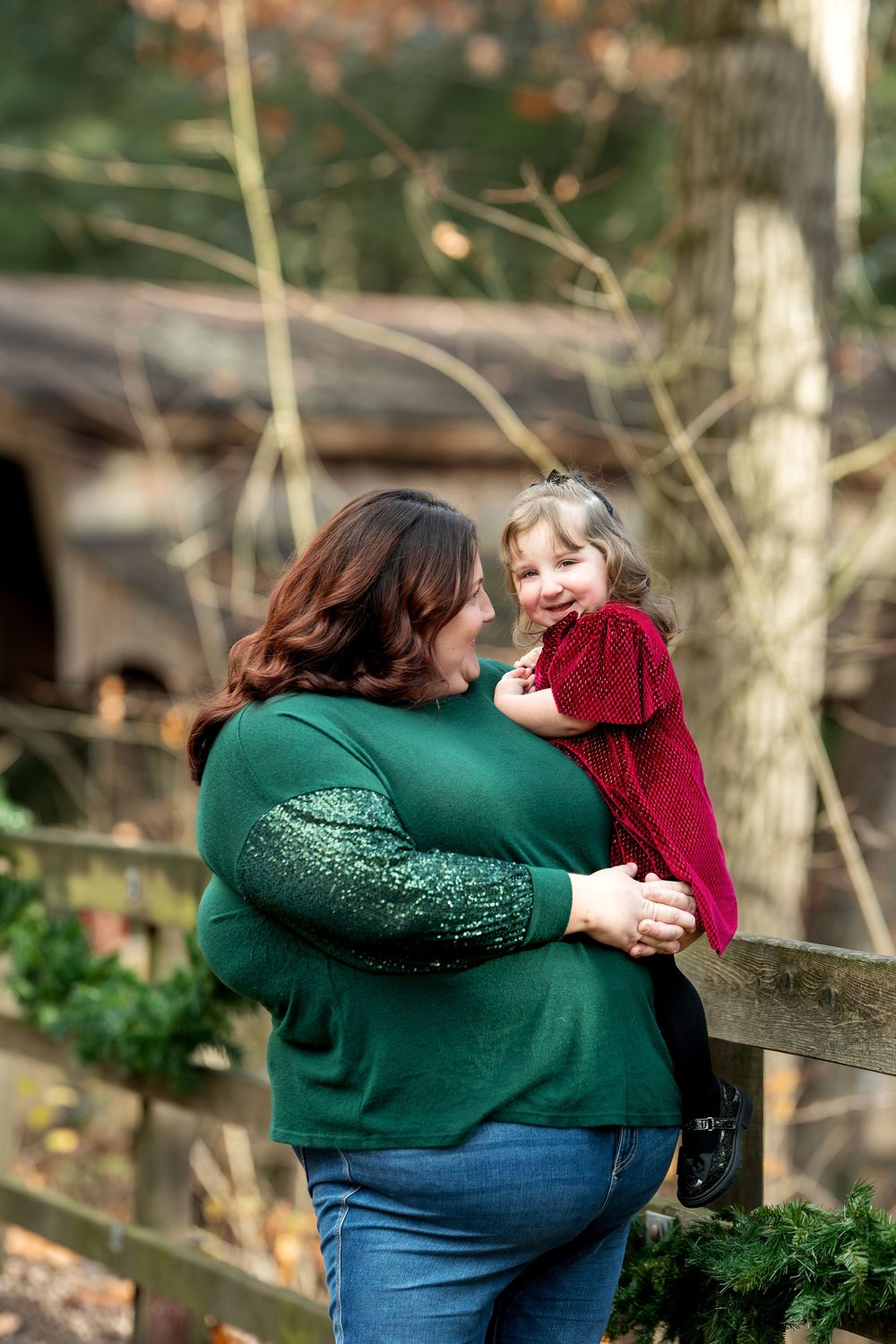 A mother in a green sequin top smiles at her laughing toddler daughter in a red dress outdoors.