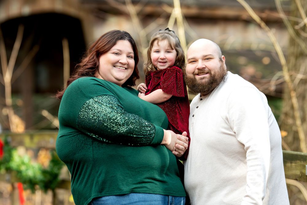 A happy family of three poses outdoors, with parents holding their young daughter in a red velvet dress.