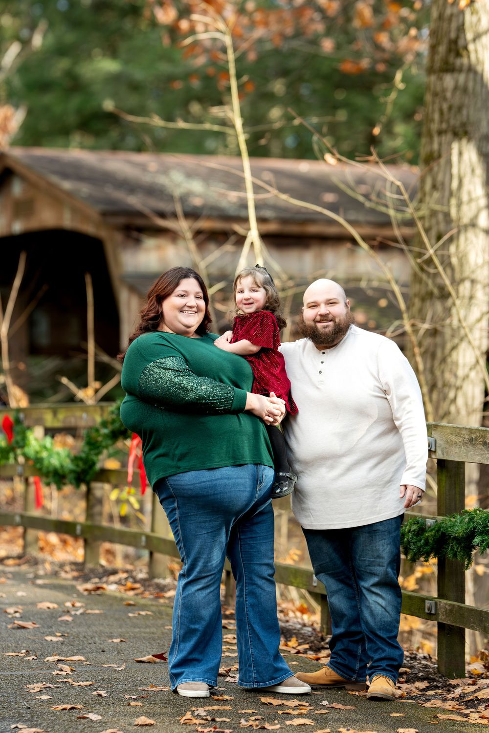 A smiling family of three stands on a leaf-covered path near a covered bridge with Christmas wreaths.