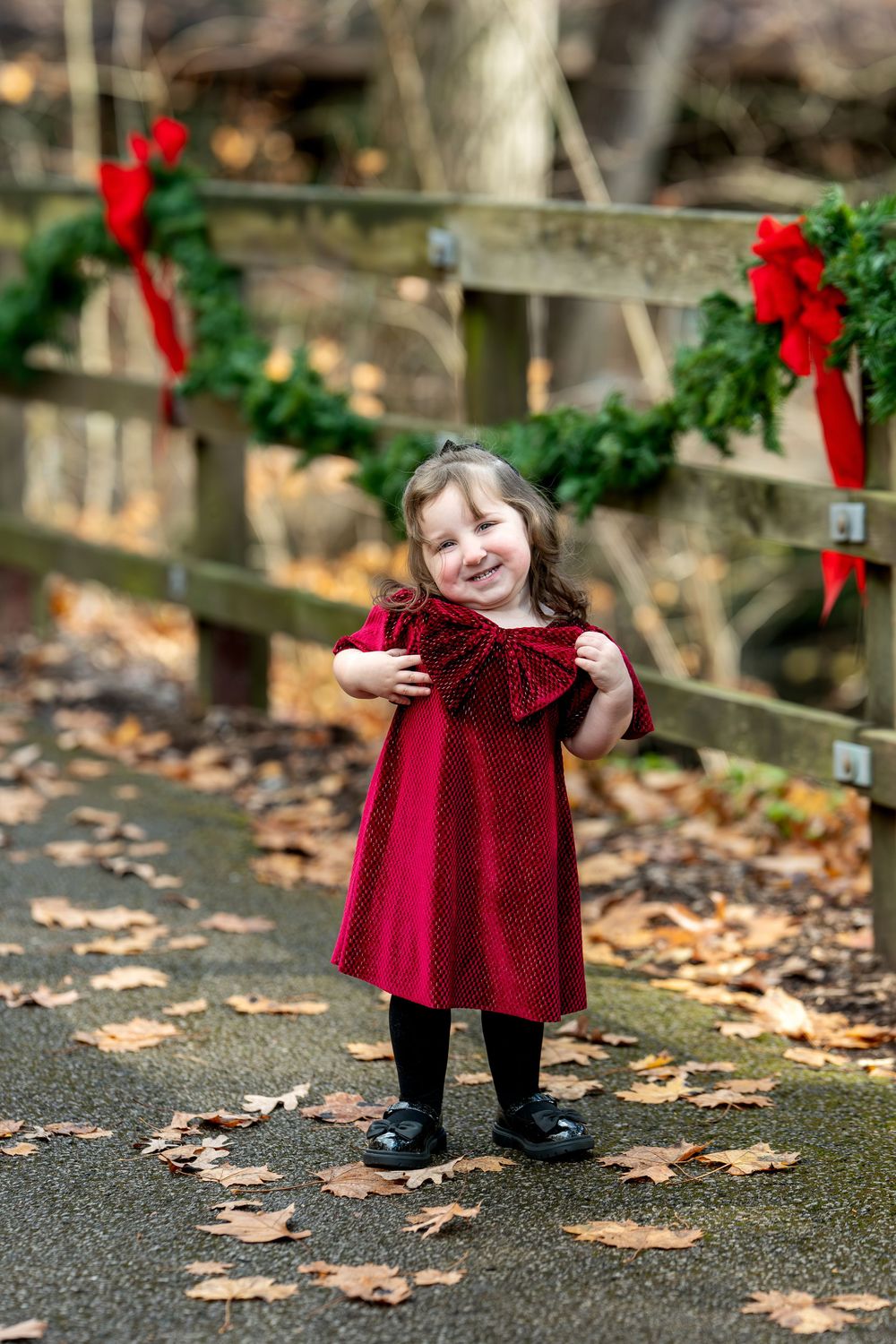 Smiling toddler girl in red holiday dress holds bow while standing by Christmas-decorated fence.