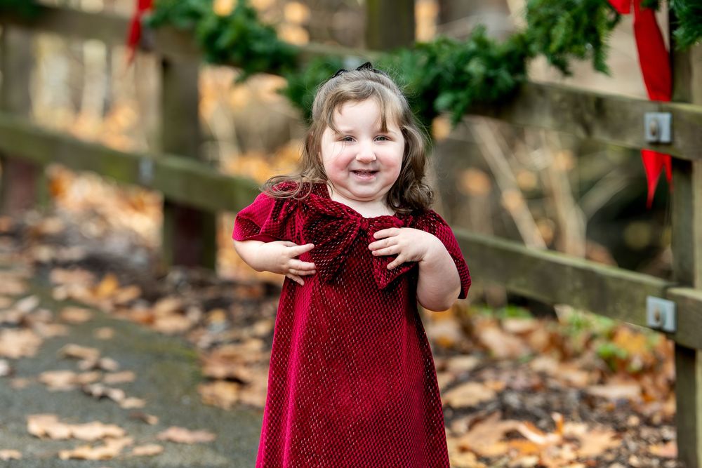 Happy little girl in red velvet dress smiles outdoors near Christmas wreaths on wooden fence.