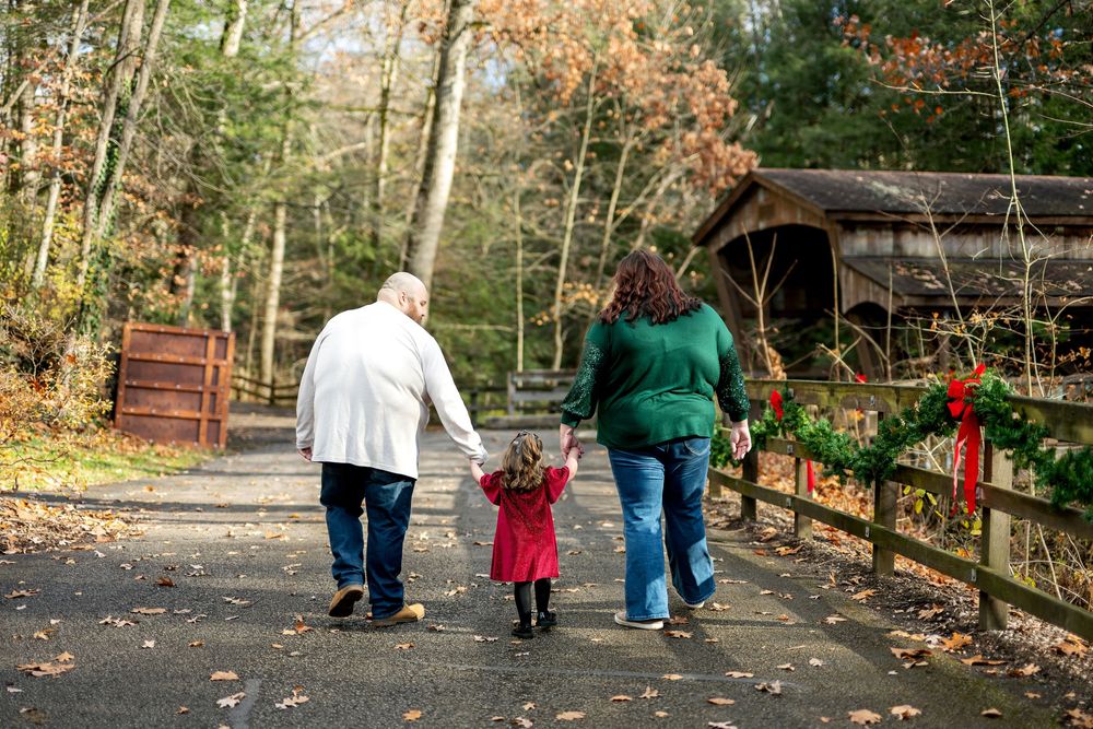 Grandparents walk hand-in-hand with toddler in red dress toward covered bridge decorated for Christmas.