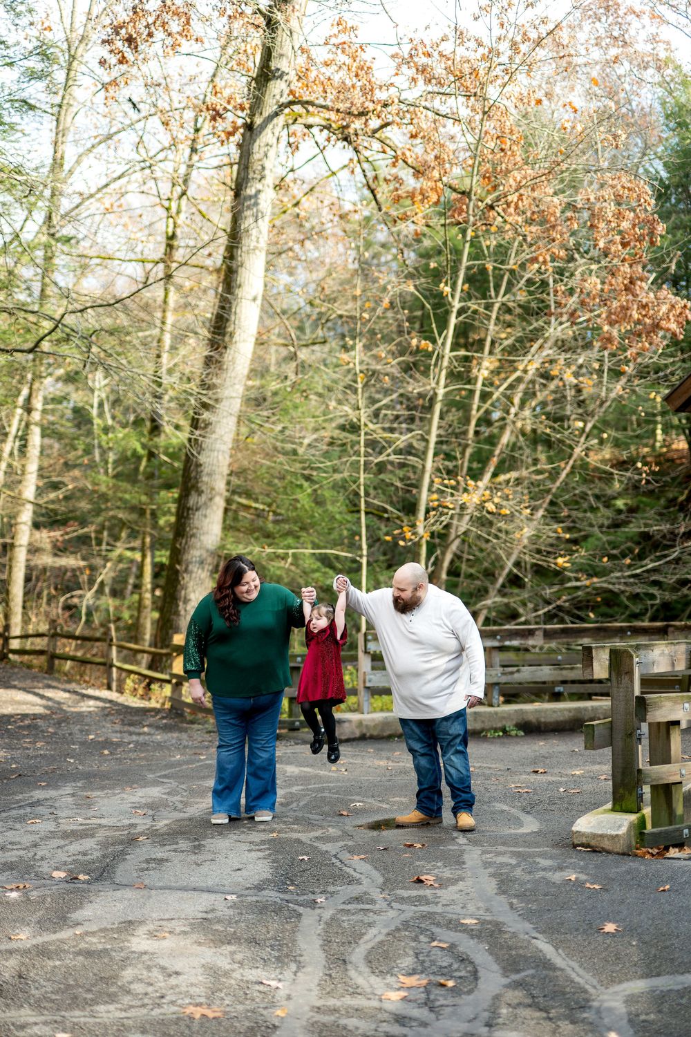Parents swing toddler girl in red dress between them on leaf-covered path in autumn park.